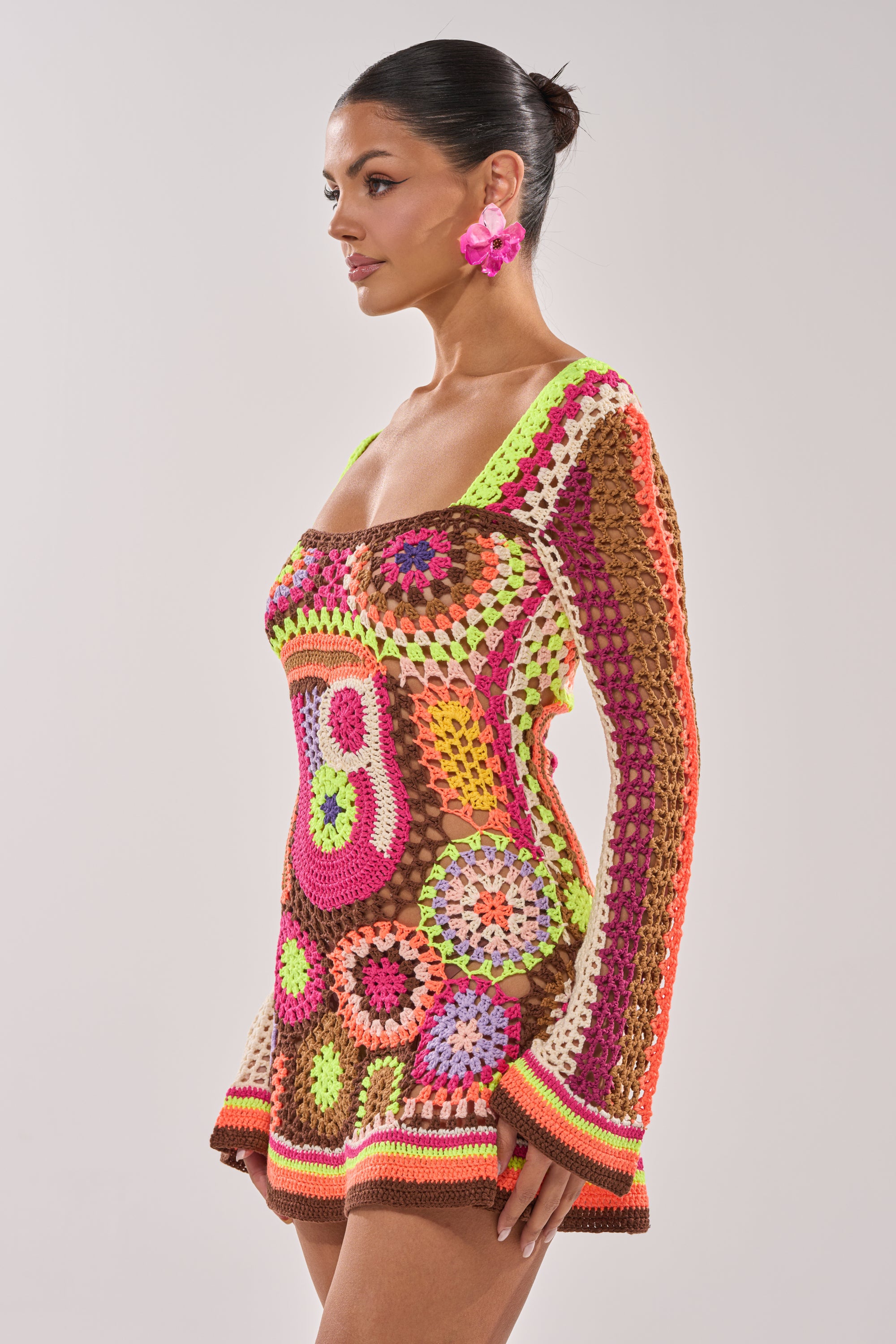 A woman with dark hair in a bun wears the CABANA CROCHET MINI DRESS, featuring colorful geometric crochet patterns, and bright pink flower earrings, standing against a plain light background. This patchwork dress brings playful summer style. 3