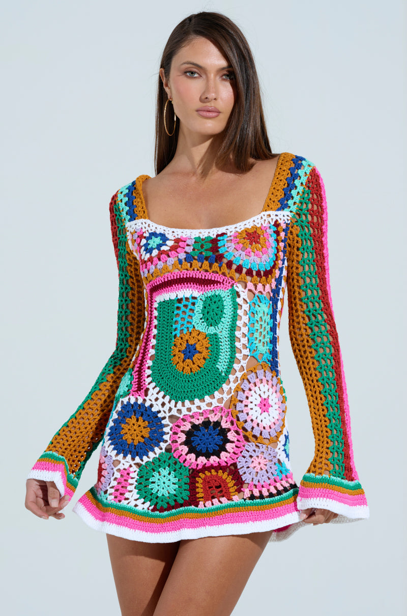 A person with long brown hair poses gracefully against a plain background in the CROCHET ALL DAY COLORBLOCK MINI DRESS, showcasing its intricate circular patterns in pink, blue, green, and orange crochet with long sleeves and a square neckline.