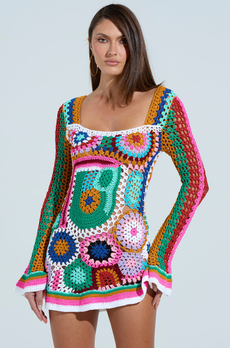 A person wearing the Crochet All Day Colorblock Mini Dress, featuring a colorful A-line silhouette with geometric patterns in vibrant pink, blue, green, orange, and white against a plain background. Long brown hair flows down as hoop earrings finish the striking look.