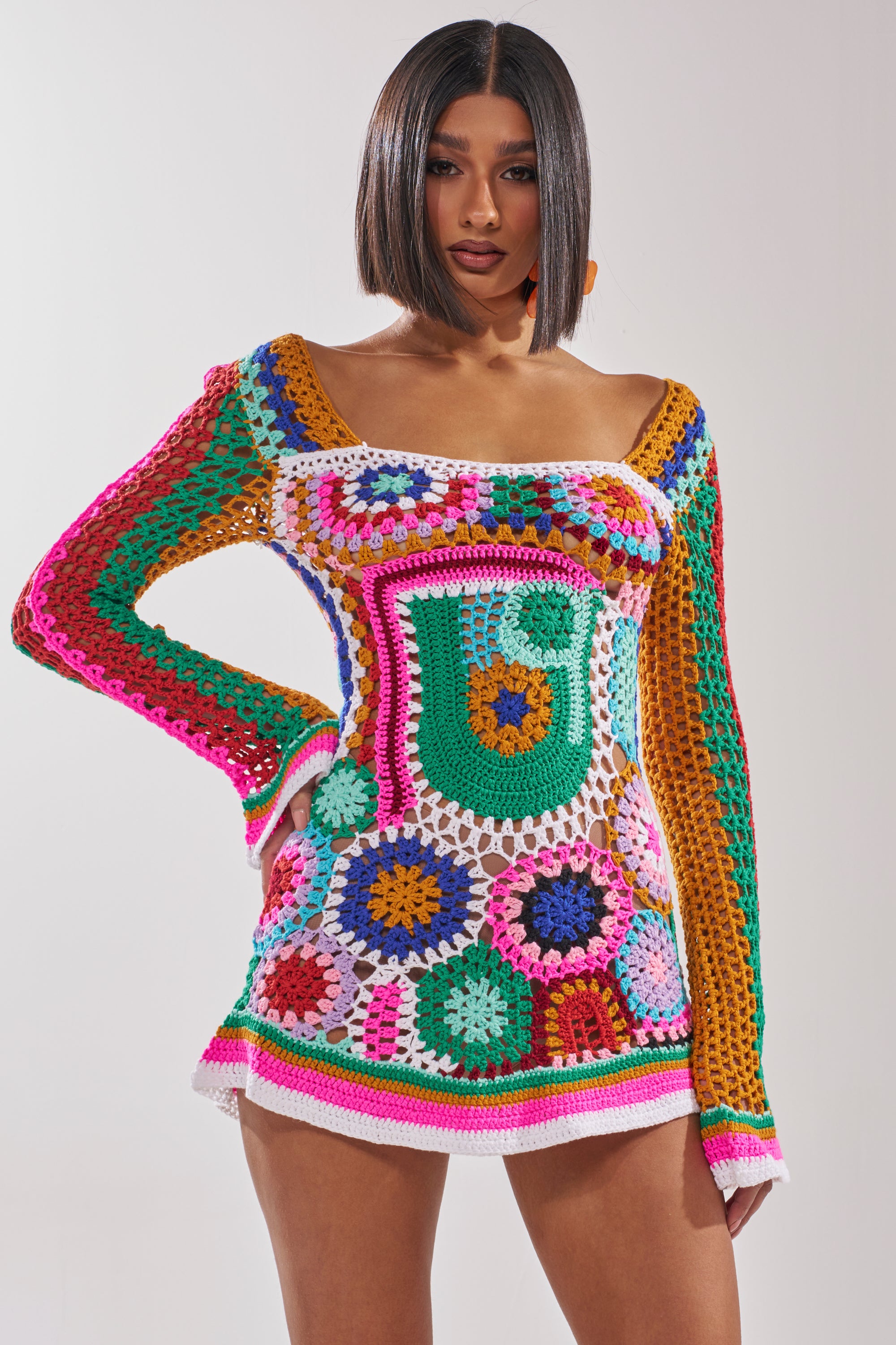 A woman poses confidently in the CROCHET ALL DAY COLORBLOCK MINI DRESS, a colorful, long-sleeved mini dress with vibrant patchwork and playful bell sleeves, making this standout piece pop against the plain background. 1