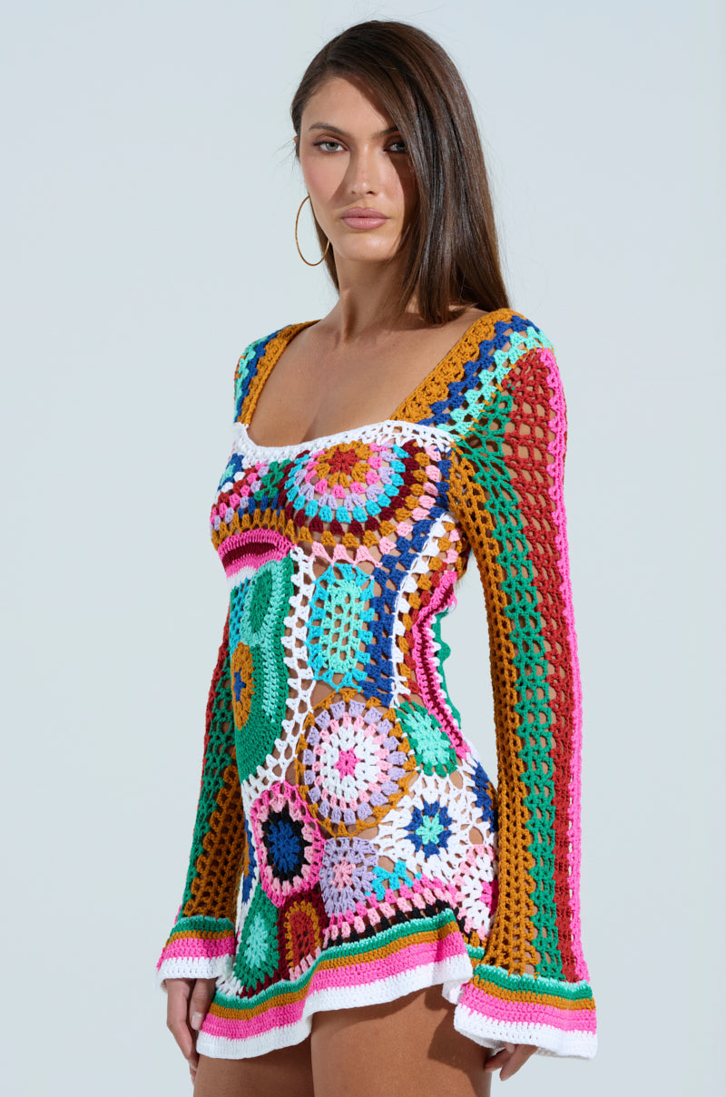 A woman confidently poses against a plain background, wearing the "Crochet All Day Colorblock Mini Dress." The colorful mini dress features long sleeves with circular patterns in pink, blue, green, mustard, and white. Bell sleeves add a playful touch. She has long brown hair.