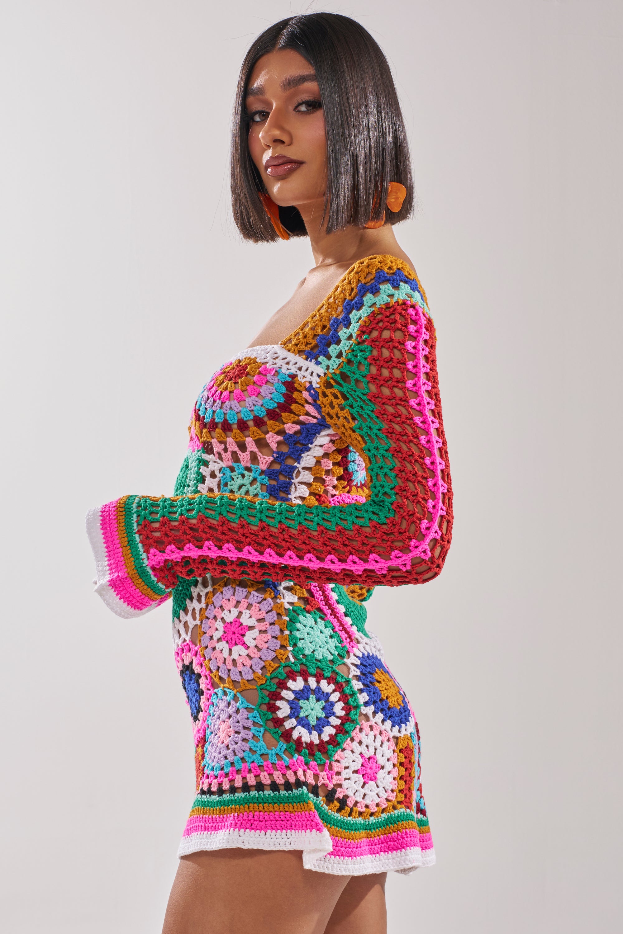A woman with straight shoulder-length hair models the CROCHET ALL DAY COLORBLOCK MINI DRESS, a vibrant mini dress with bell sleeves and multicolored circular patterns, set against a plain light background. 3