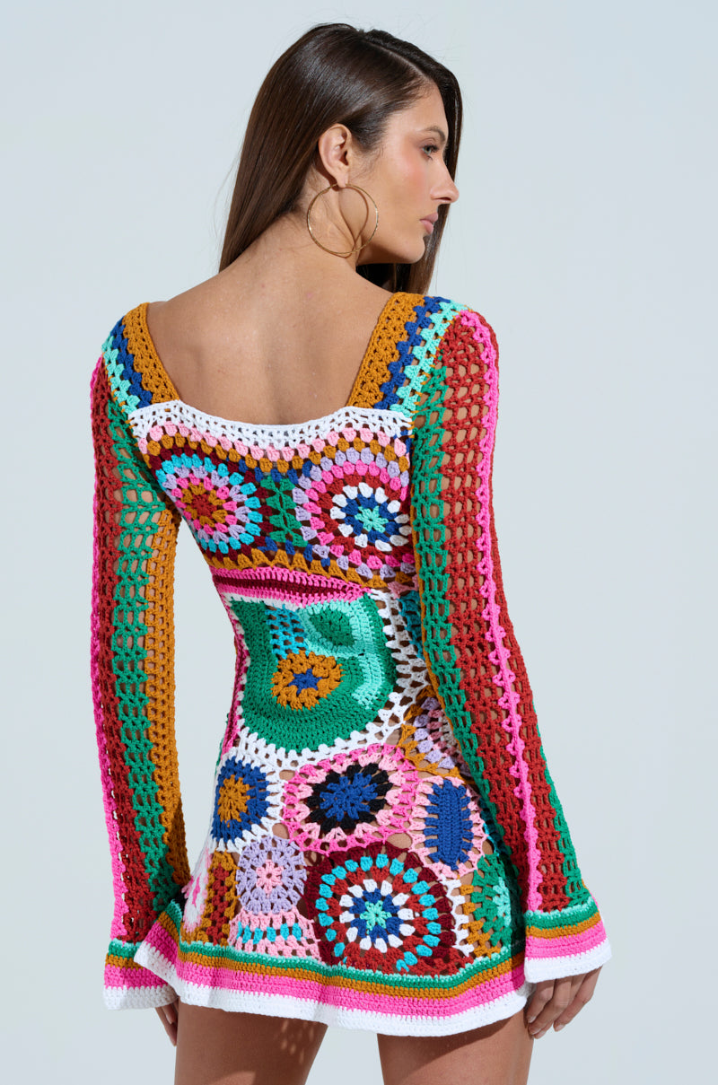 A woman with long brown hair models the "Crochet All Day Colorblock Mini Dress." Featuring intricate circular patterns in vibrant pink, green, blue, yellow, and orange hues with an A-line silhouette, she poses against a plain background highlighting the dress's back and sides.