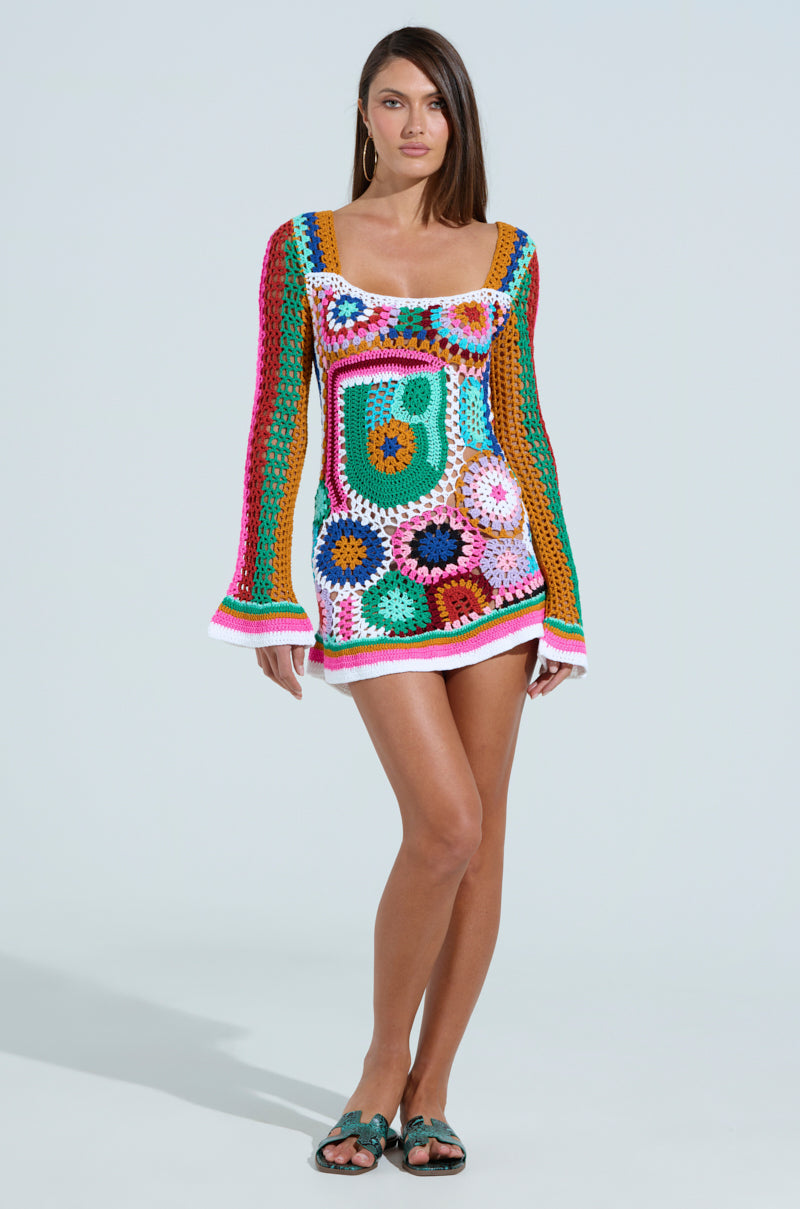A woman in a CROCHET ALL DAY COLORBLOCK MINI DRESS stands against a plain background. The vibrant A-line mini features bright colors like pink, green, and orange in various patterns. She has long hair and wears sandals.