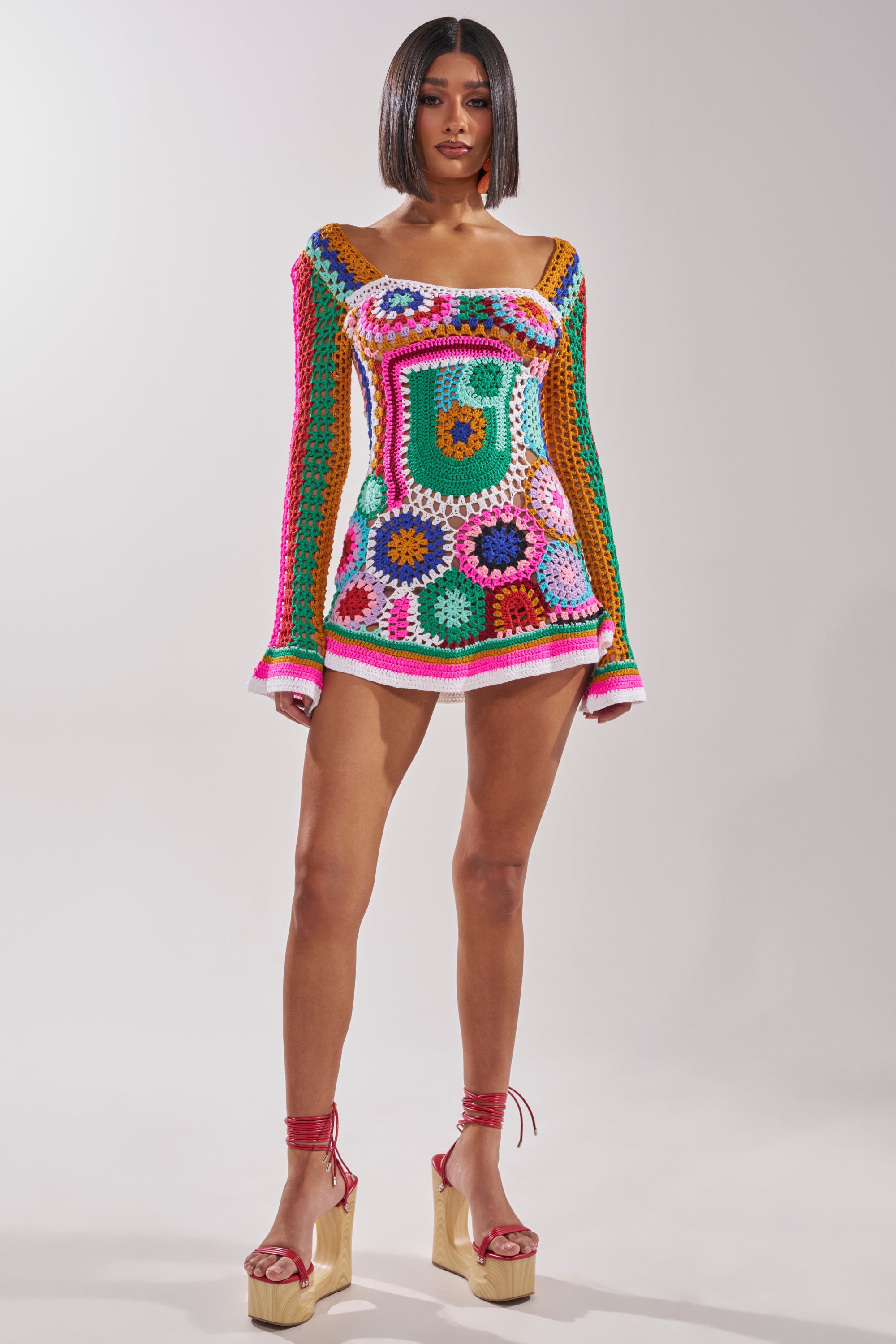 A woman stands confidently in the CROCHET ALL DAY COLORBLOCK MINI DRESS, featuring vibrant colorblock crochet patterns and subtle bell sleeves, paired with red tie-up platform sandals against a plain light background. 5