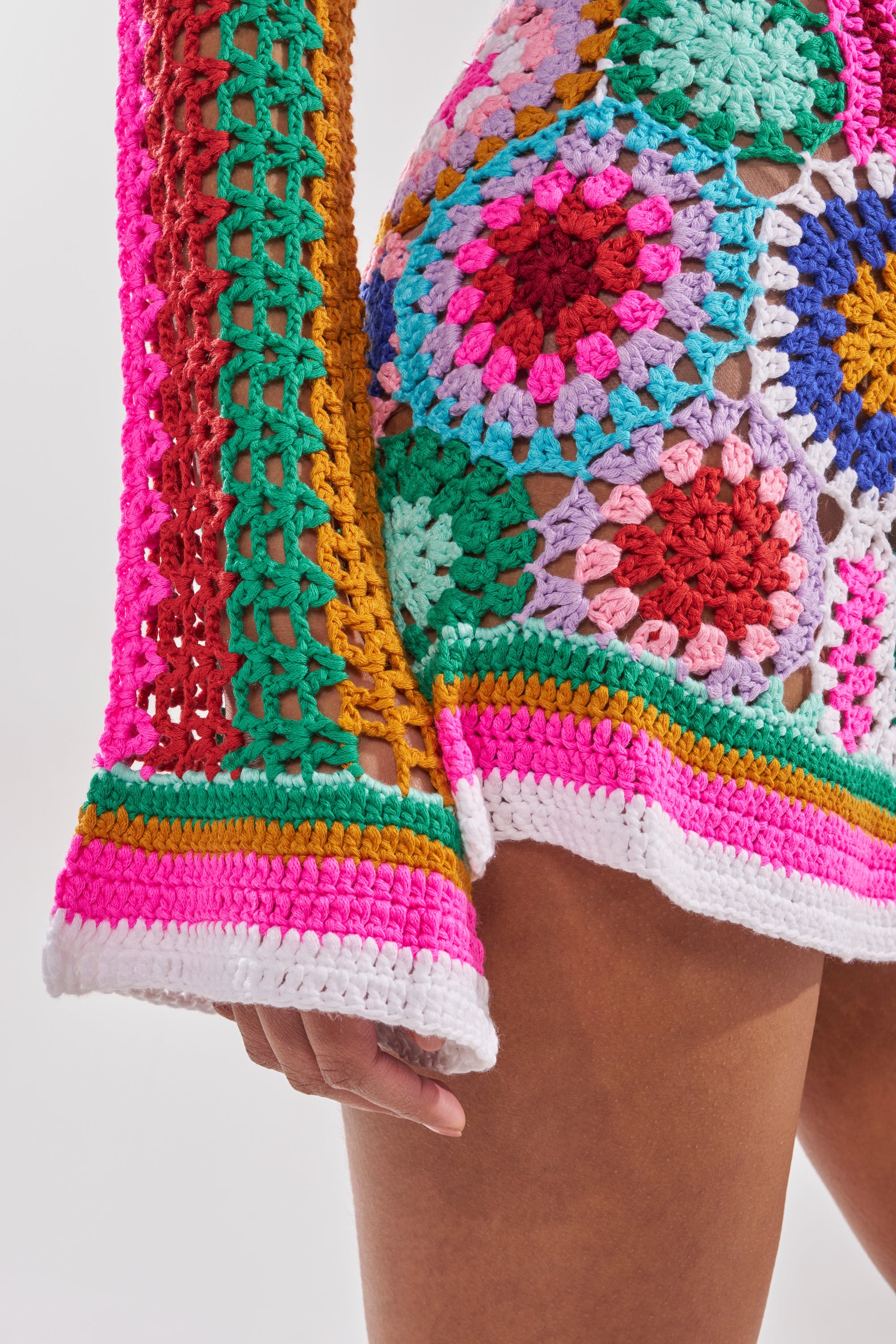 A close-up of a person wearing the CROCHET ALL DAY COLORBLOCK MINI DRESS, featuring colorful geometric and floral crochet patterns in red, green, yellow, pink, blue, and white. Their hand rests by their side. 8