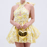 A woman wears THE TOPIC AND TOP PICK BROCADE MINI DRESS IN YELLOW, featuring a halter neckline and large back bow, while holding a shiny gold heart-shaped handbag against a plain light background.