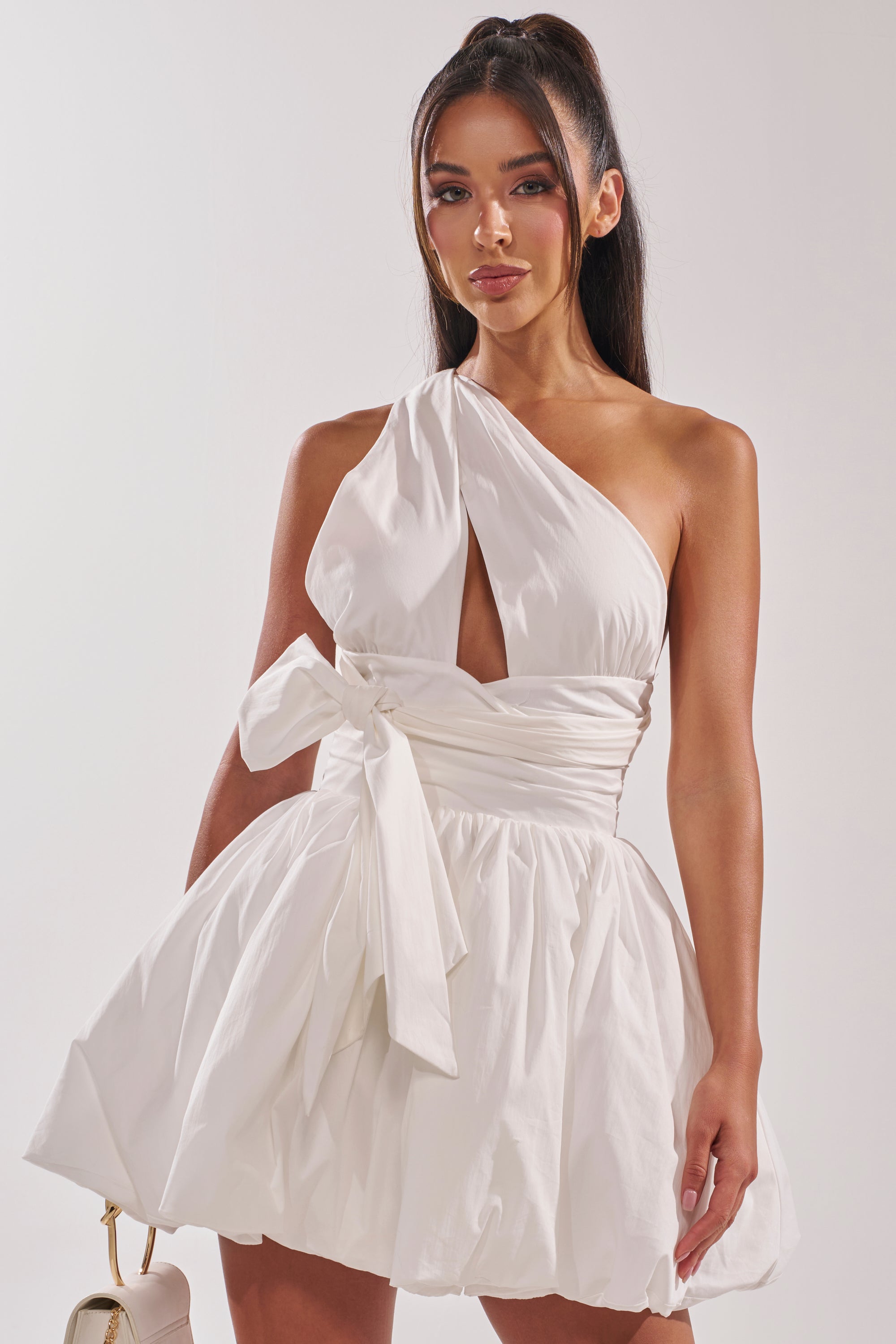 A woman stands against a plain light background, holding a small handbag and wearing the PRETTY IN POPLIN STRAPPY MULTIFUNCTION MINI DRESS IN WHITE featuring a front cutout, waist bow, and flared skirt.
