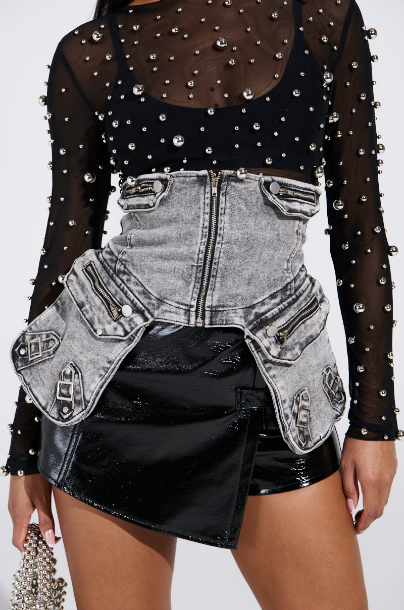 Wearing a black mesh top with pearls and a gray corset with cargo pockets and zippers, they pair it with a shiny black asymmetrical skirt. They hold the Maeve Embellished Stretch Denim Belt as a pearl-studded purse against a plain background.