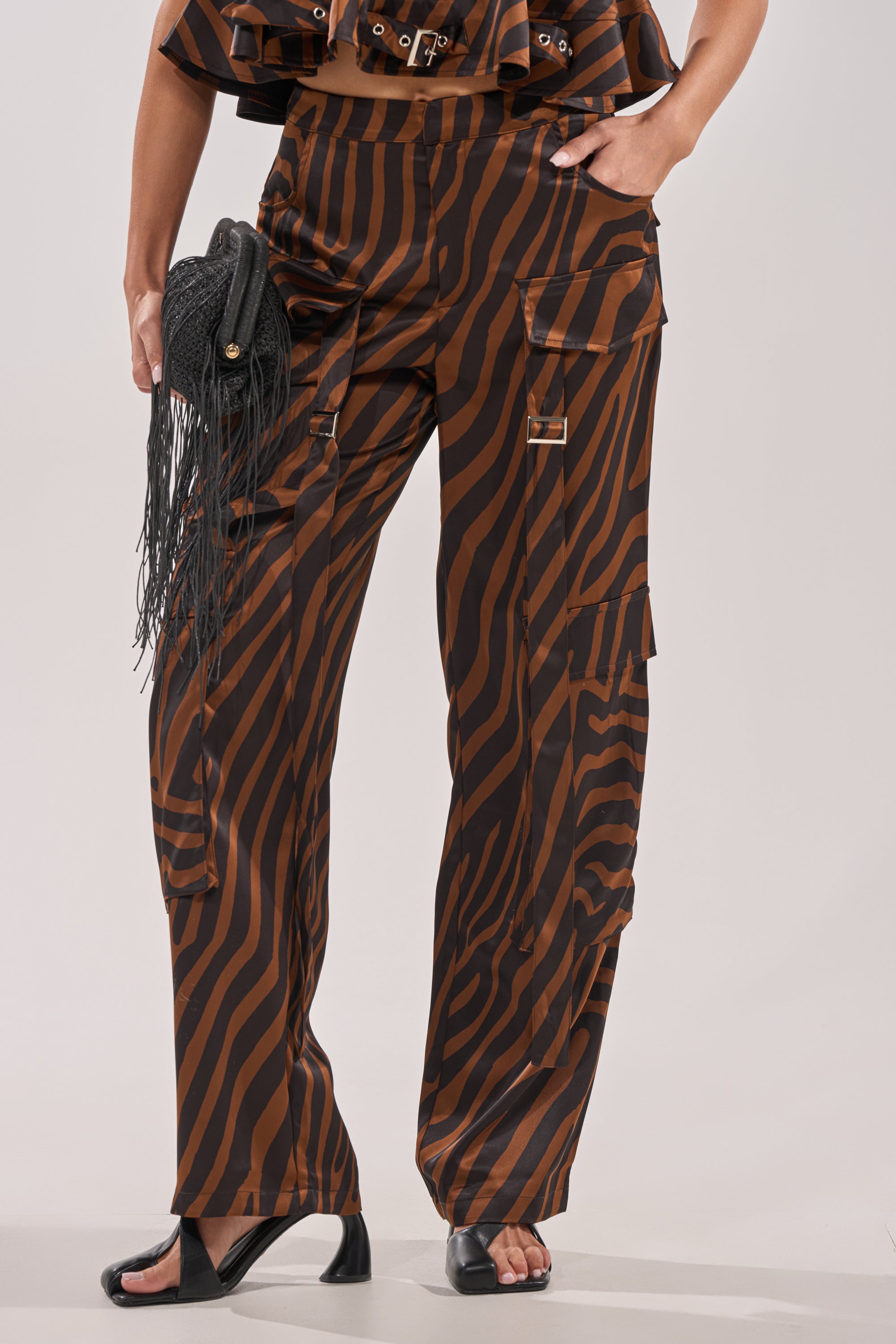 A person wearing INTO THE WILD ZEBRA TROUSER with cargo pockets, black open-toe heels, and holding a black woven fringe clutch. Only the lower body is visible. 1