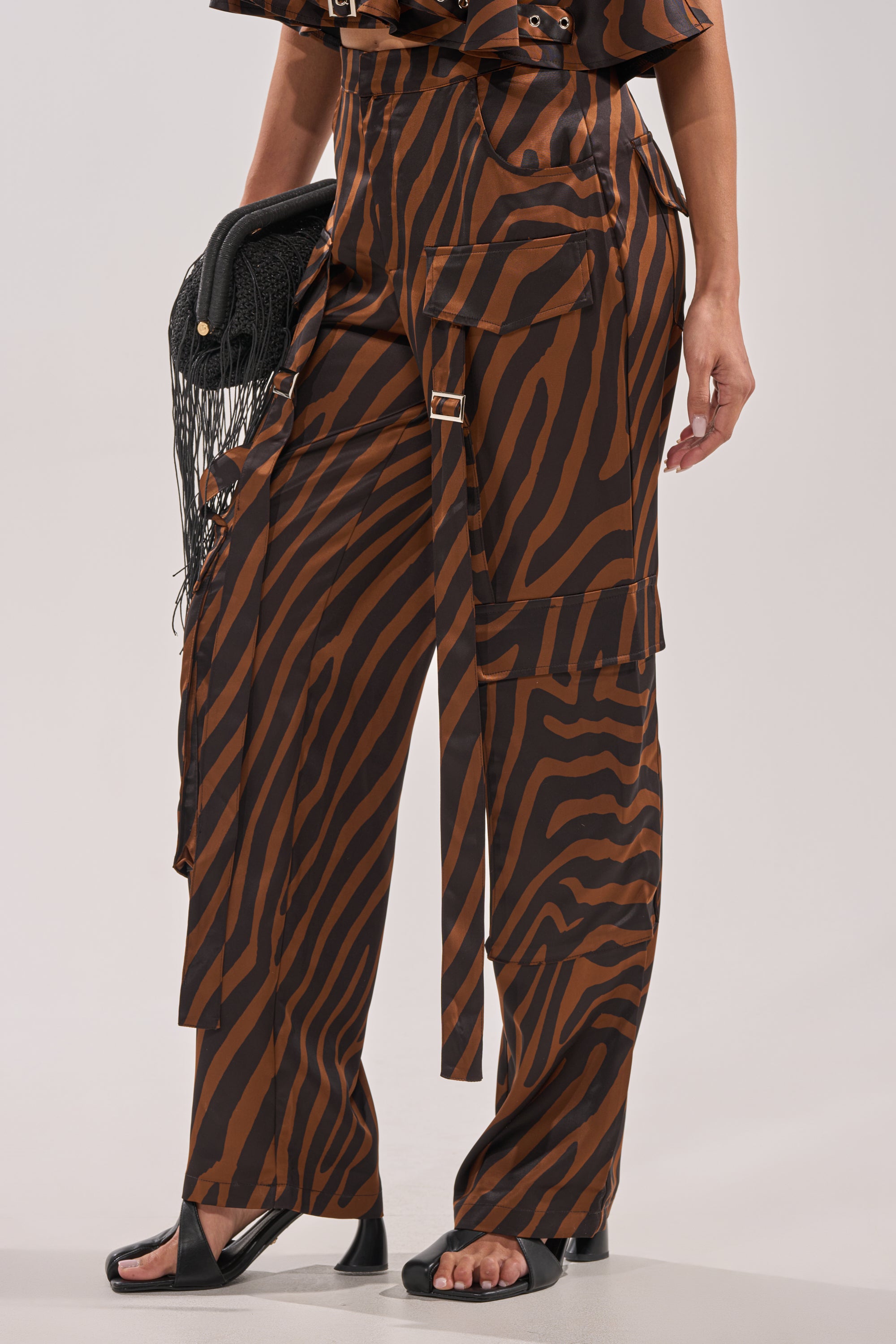 Wearing the INTO THE WILD ZEBRA TROUSER—brown and black zebra print cargo-style pants with extra straps—paired with black sandals and a black fringed clutch. Only the lower half of the outfit is shown. 3