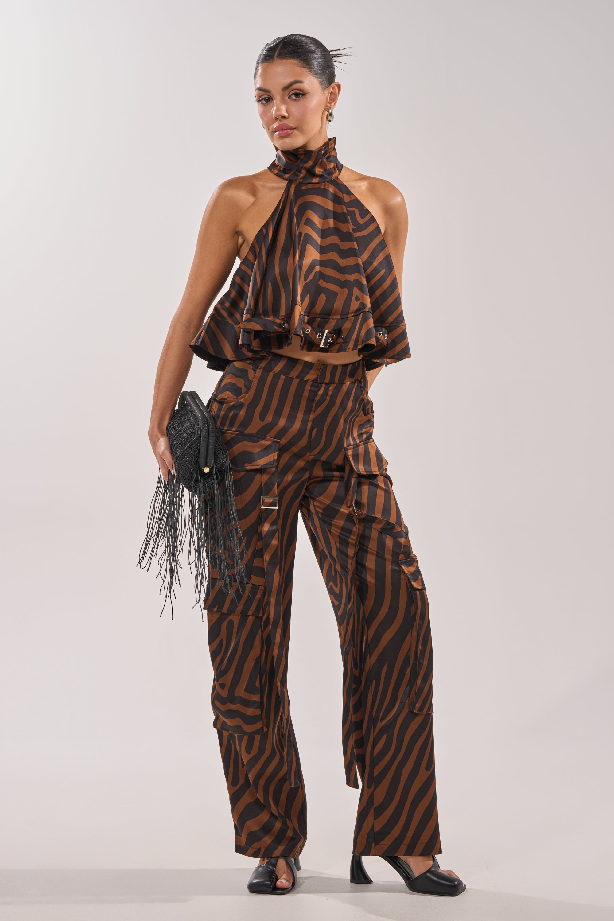 A woman in INTO THE WILD ZEBRA TROUSER and a matching halter top stands confidently, holding a black fringed clutch. She wears black sandals, has slicked-back hair, and poses stylishly against a plain background. 7
