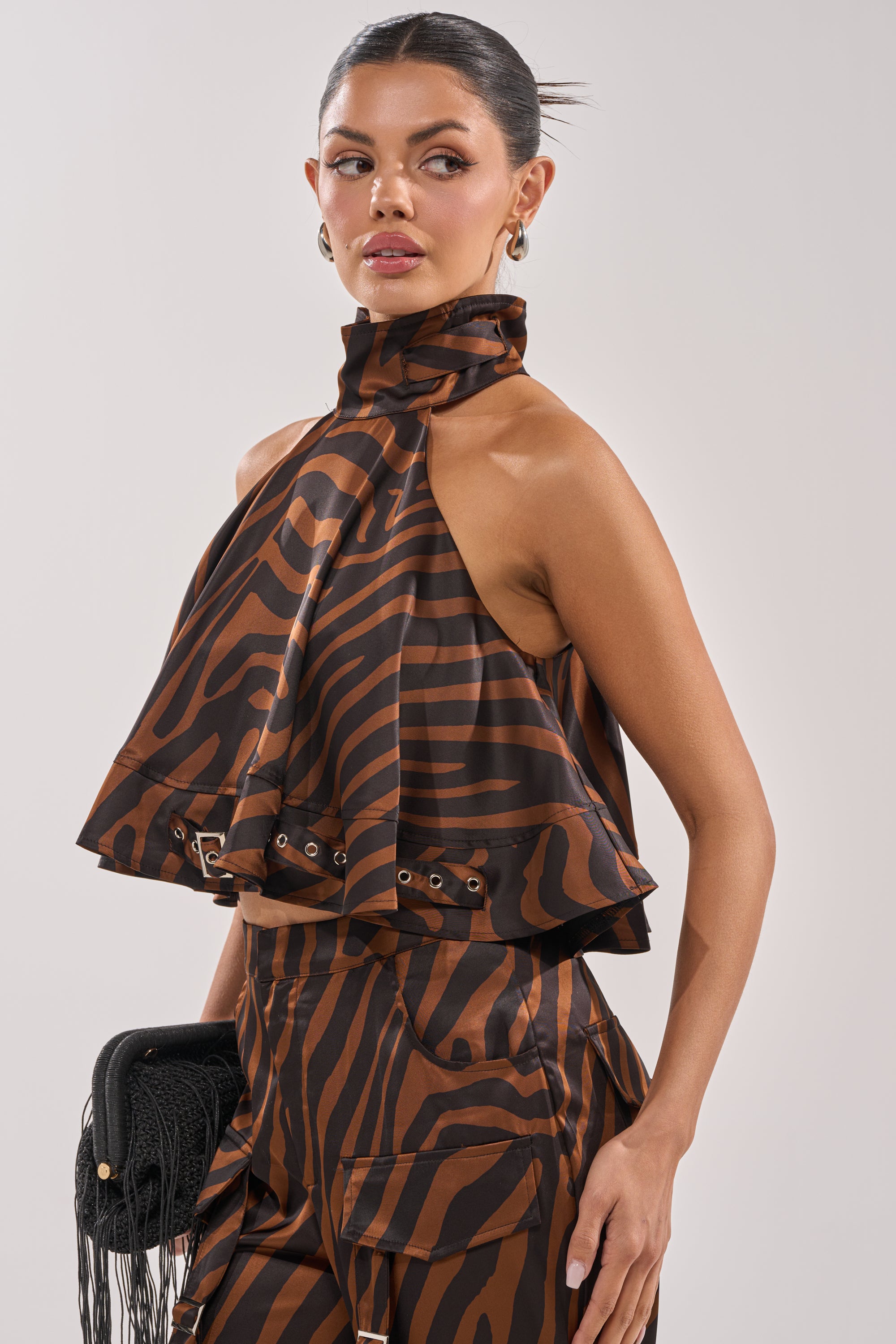 A woman in the INTO THE WILD ZEBRA HALTER NECK BLOUSE and matching high-waisted pants stands with a black fringe clutch, looking to the side. Her hair is slicked back and she wears gold hoop earrings. 3