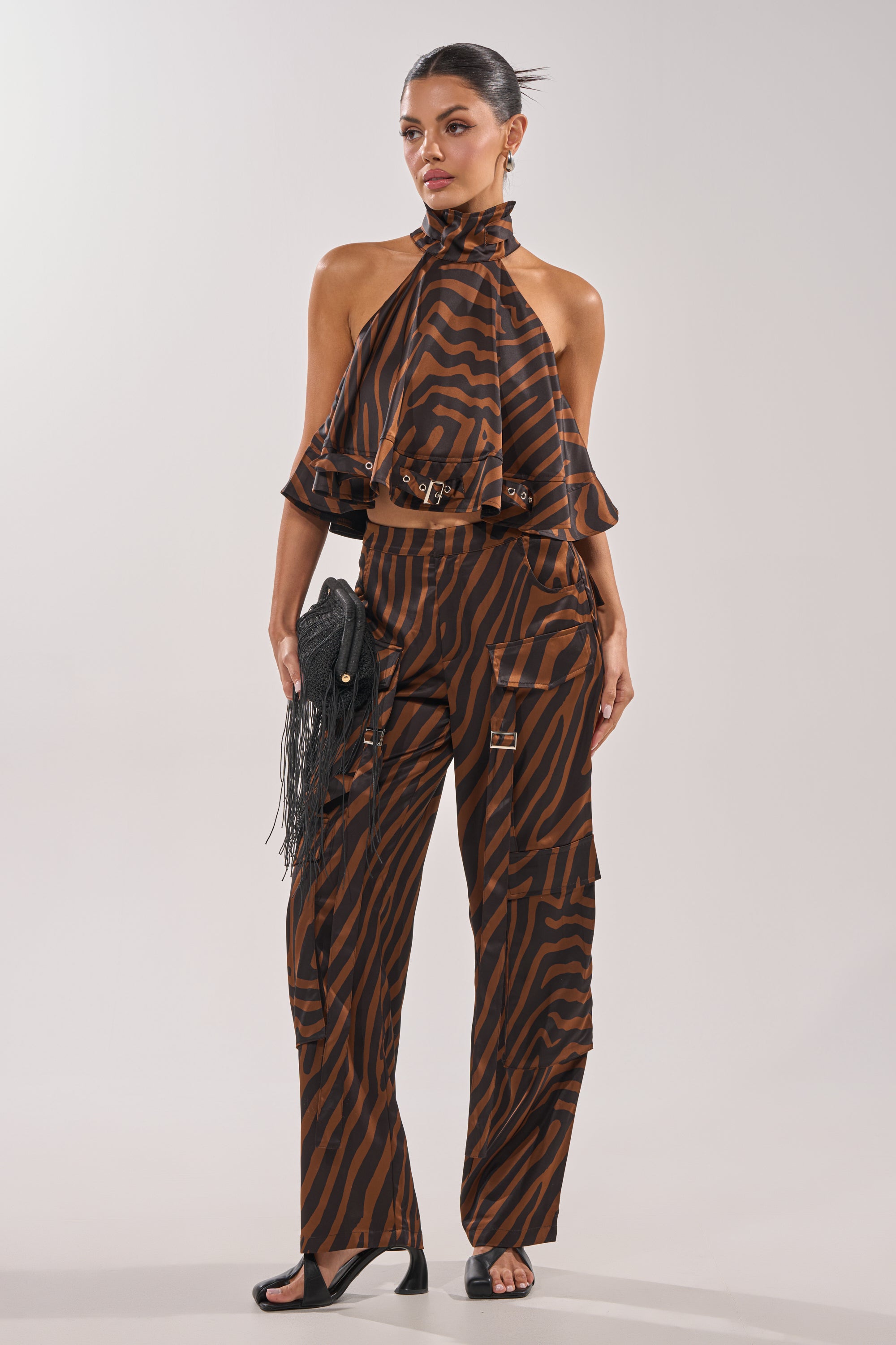 A woman wears the INTO THE WILD ZEBRA HALTER NECK BLOUSE with matching wide-leg pants, accessorized with a black fringe clutch and black heeled sandals, standing against a plain light background. 5
