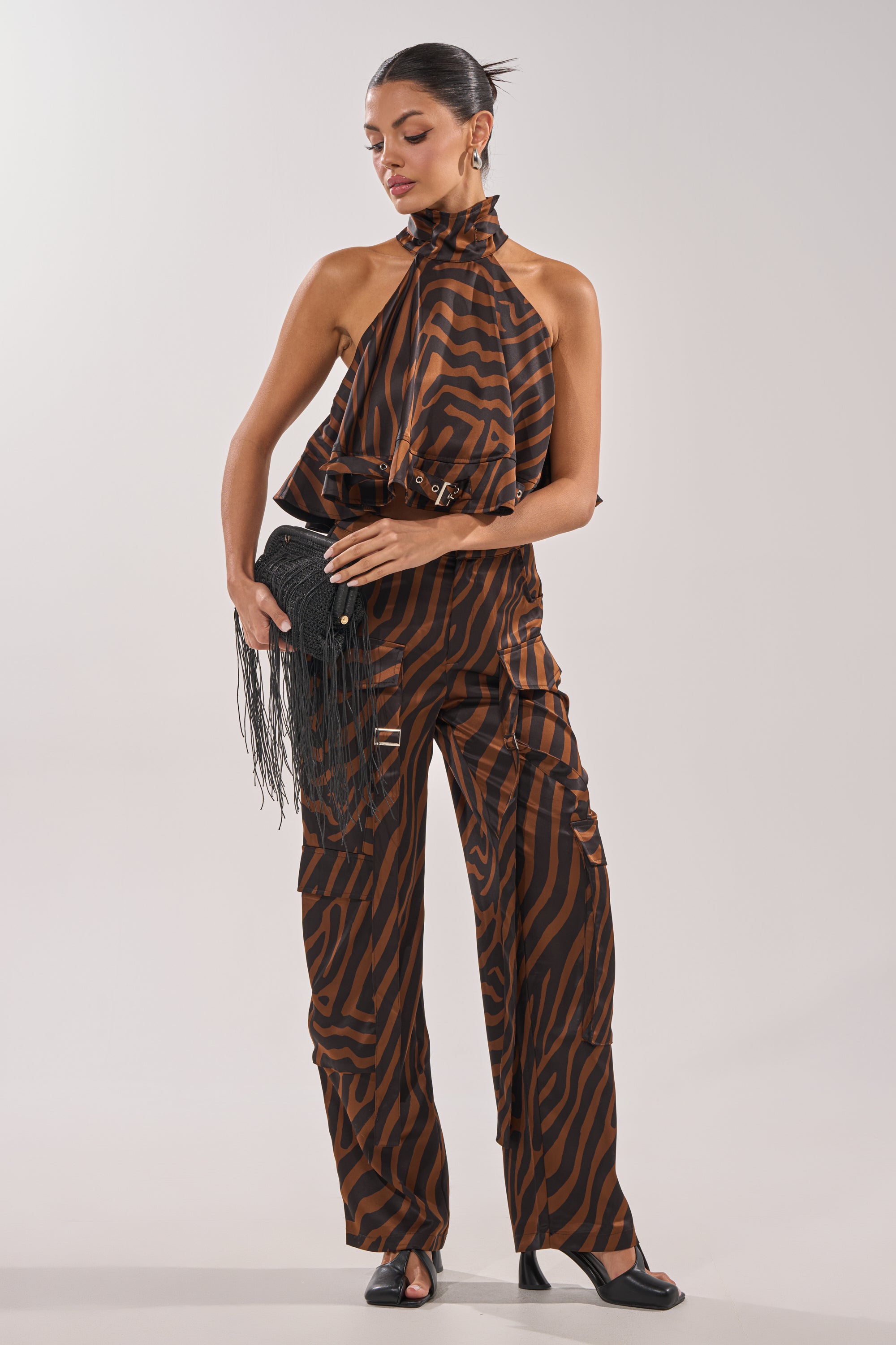 A woman poses in the INTO THE WILD ZEBRA HALTER NECK BLOUSE with matching wide-leg pants, holding a black fringed clutch. She wears black heels and a sleek bun, highlighting her party-ready zebra print look. 6