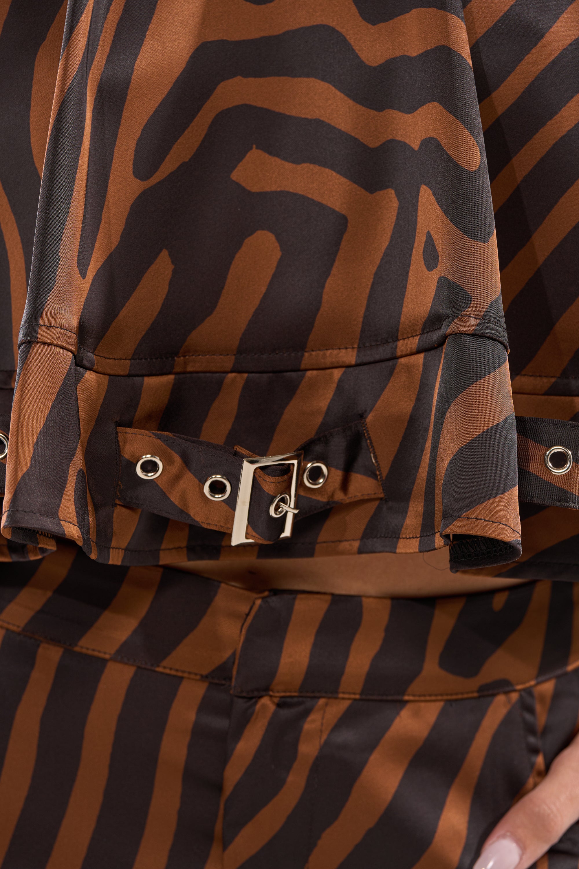 Close-up of the INTO THE WILD ZEBRA HALTER NECK BLOUSE in satin with brown and black zebra stripes, silver buckle and grommets. A manicured hand appears on the right. 8