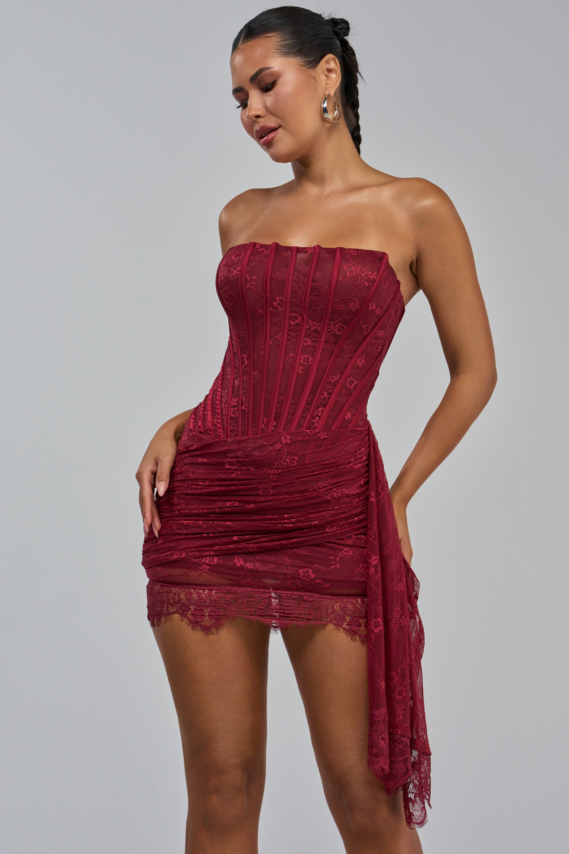 A woman models the SABRINA LACE LAYERED MINI DRESS IN BURGUNDY—a strapless, form-fitting mini dress featuring intricate lace layers, a ruched skirt, and a sophisticated side drape—paired with hoop earrings as she poses against a plain gray background.