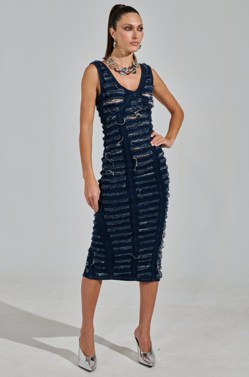 A woman wears the SHE'S GOT IT SHREDDED Denim Midi Dress in Dark Blue, posing confidently with one hand on her hip. She accessorizes with silver high heels and a chunky necklace, and her hair is styled in a sleek ponytail against a light, plain background.