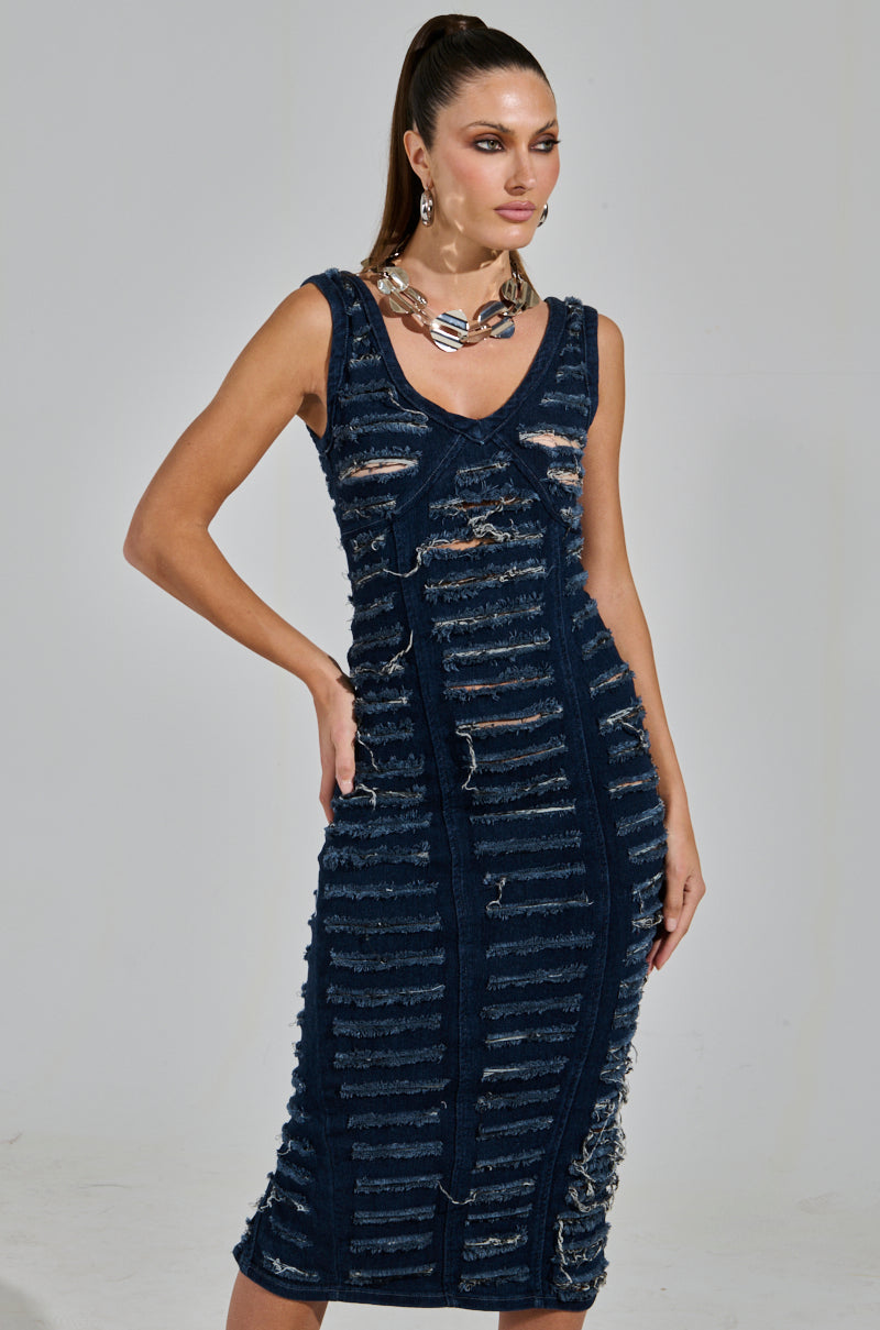 A woman poses in the SHE'S GOT IT SHREDDED DENIM MIDI DRESS IN DARK BLUE, featuring curve-hugging distressed denim with shredded stripes. She accessorizes with chunky metallic jewelry and a high ponytail against a neutral gray backdrop, highlighting her edgy style.