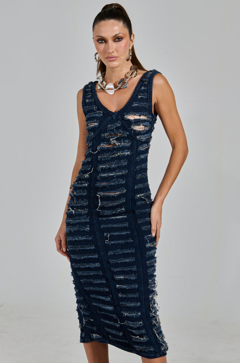 A person models the "SHE'S GOT IT SHREDDED DENIM MIDI DRESS IN DARK BLUE," featuring a sleeveless, knee-length design with frayed lines. With their hair pulled back, they accessorize with a chunky necklace and hoop earrings against a plain background.