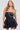A woman exudes confidence as she wears the FELT CUTE POPLIN MINI DRESS IN BLUE PINSTRIPE, characterized by its strapless design and voluminous skirt. Layered pearl necklaces and thigh-high white lace stockings complement her poised expression against a plain background.