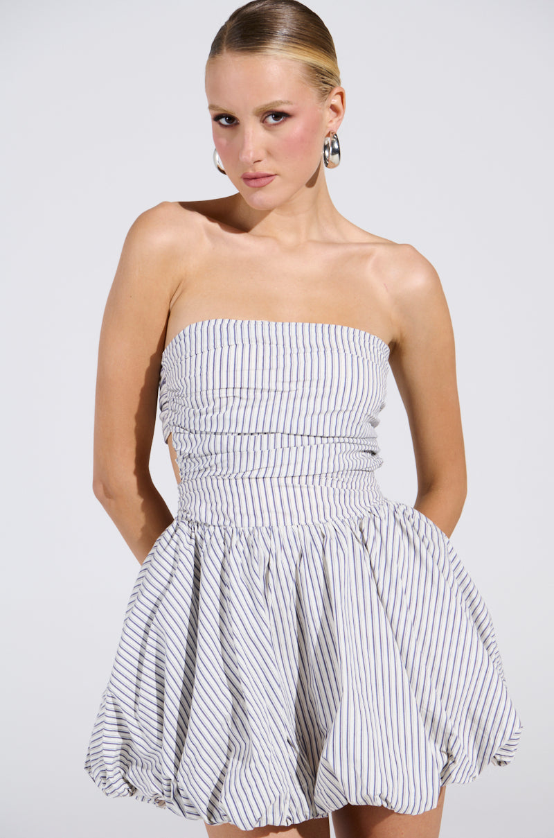 Wearing the "A Stroll Down Soho Pinstriped Mini Dress," with a strapless neckline and flared skirt, someone showcases urban-chic style. Thin black stripes enhance the look, completed by slicked-back hair and large hoop earrings against a plain white backdrop.
