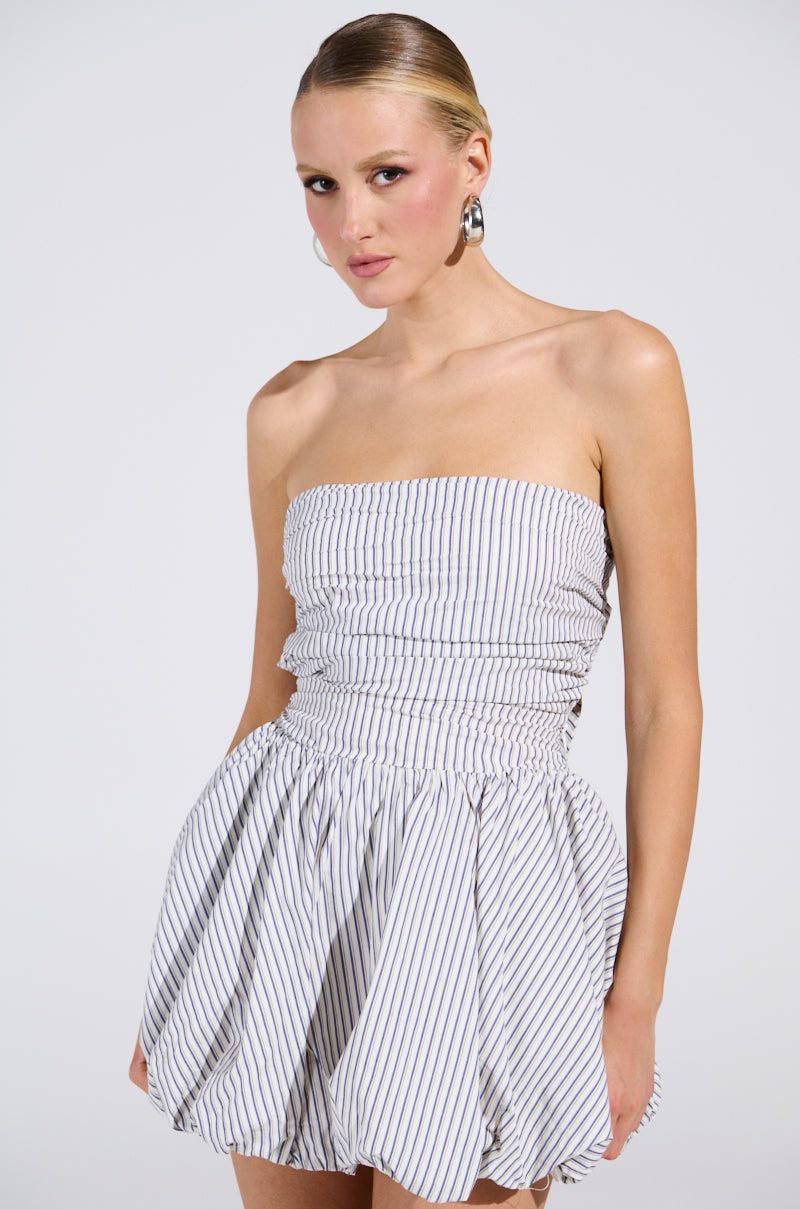 In the A Stroll Down Soho Pinstriped Mini Dress, a woman exudes urban-chic elegance with its strapless design, gathered waist, and balloon skirt. Her hair is pulled back as she poses against a plain white background.