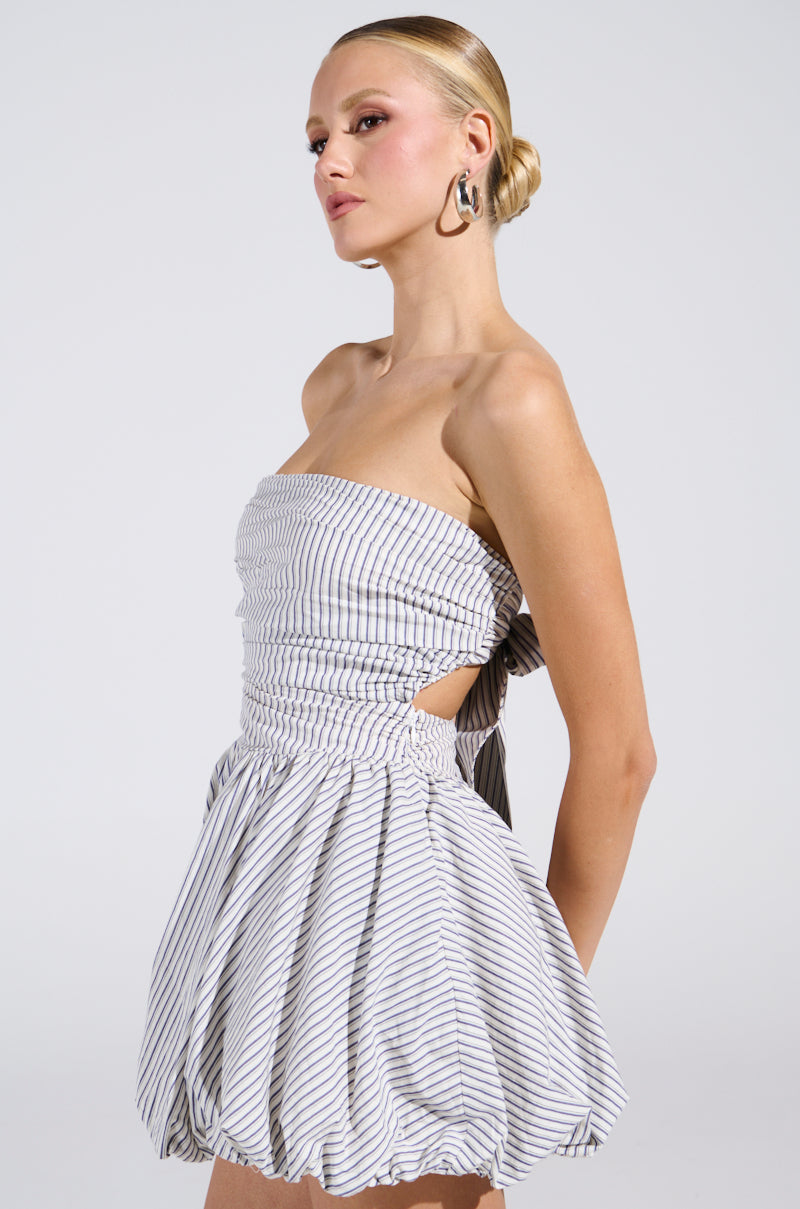 Person in "A Stroll Down Soho" pinstriped mini dress with bubble skirt, looking to the side. Their hair is styled in a sleek bun, adorned with hoop earrings against a neutral studio backdrop.