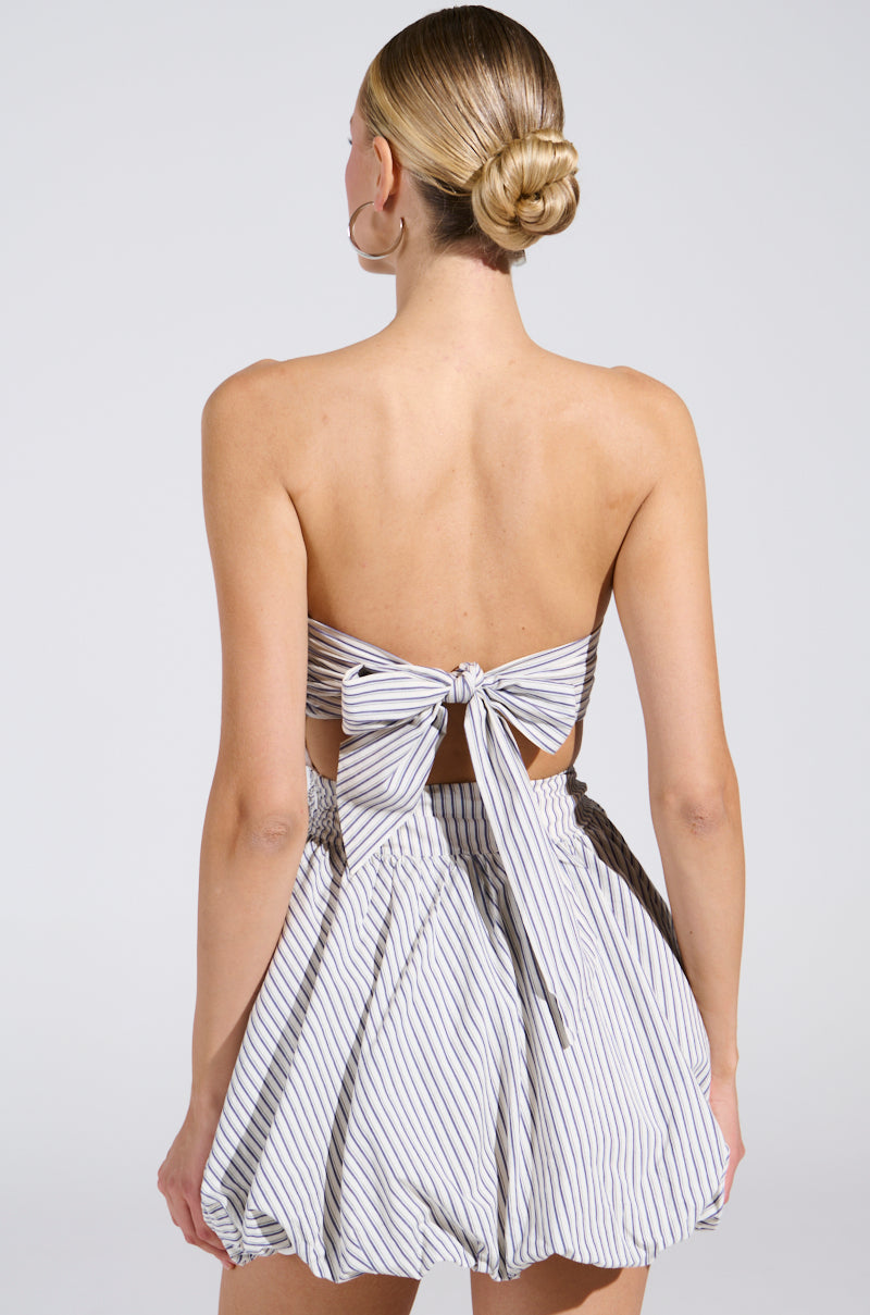 A person with blonde hair in a bun is seen from behind wearing the A STROLL DOWN SOHO PINSTRIPED MINI DRESS, featuring an urban-chic, striped backless design with a large bow and voluminous skirt. The background is plain and light-colored.