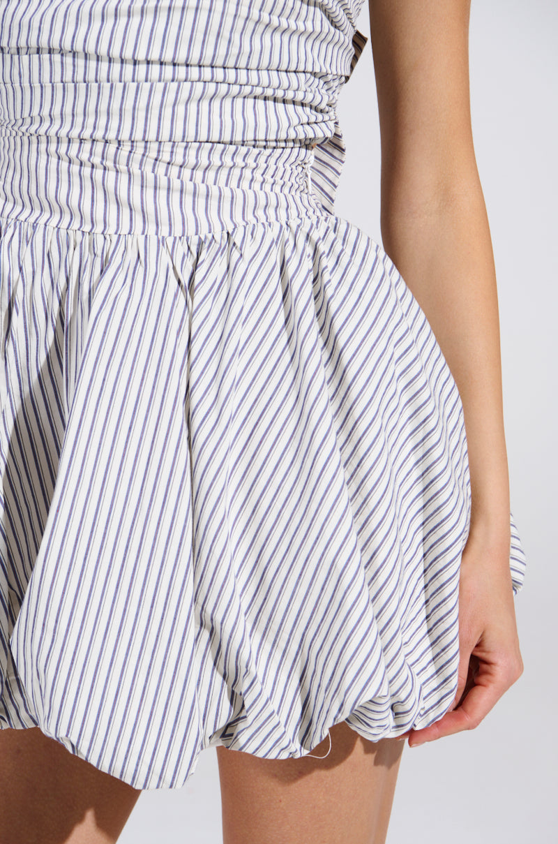 Wearing the A STROLL DOWN SOHO PINSTRIPED MINI DRESS featuring blue and white vertical stripes, a fitted waist, and flared skirt frames the visible arm on the right against a plain backdrop.