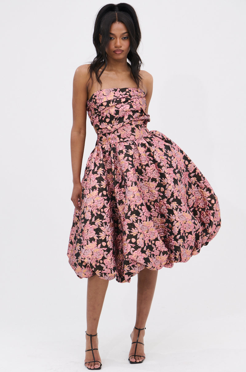 A woman wears the CHARLOTTE BROCADE MIDI DRESS featuring a ruched bodice and bold pink florals on black, styled with strappy black heels and loose wavy hair for a chic look.