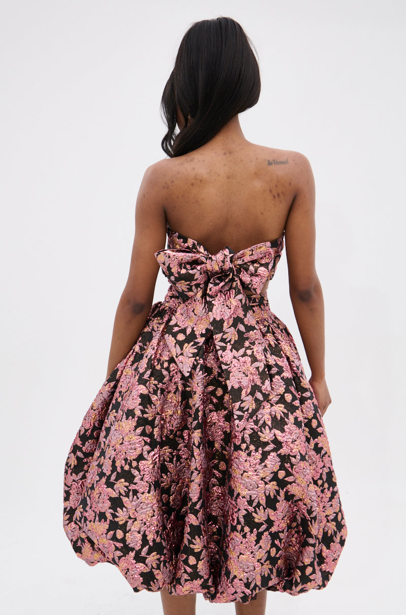 A woman with long dark hair, facing away against a white background, wears the CHARLOTTE BROCADE MIDI DRESS—a strapless black midi dress with metallic pink and gold floral brocade, ruched bodice, voluminous skirt, and an oversized back bow.