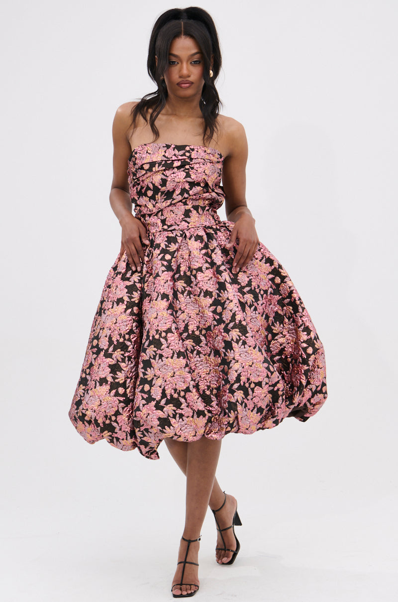 A woman models the CHARLOTTE BROCADE MIDI DRESS with a ruched bodice and pink floral pattern on dark fabric. She stands against a white backdrop, one leg forward and hands holding the skirt sides.