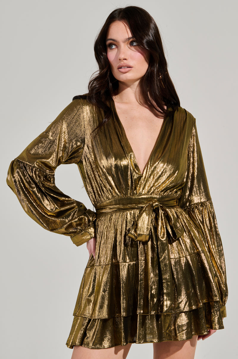 A woman stands against a plain background, wearing the GLOW BABY METALLIC MINI DRESS. The dress showcases its shiny metallic gold fabric and tiered ruffled skirt, complemented by a plunging V-neckline and a tied waist. Her long, dark hair adds to the elegance of her pose.