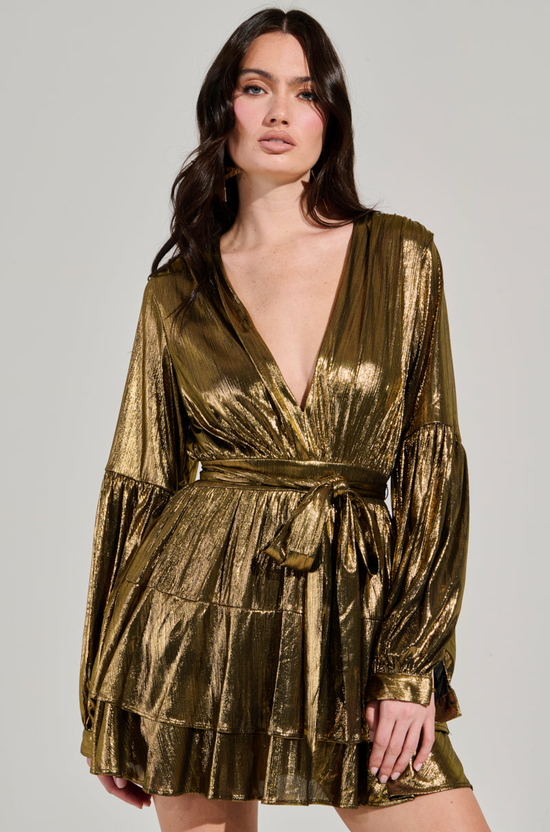 A woman with long dark hair confidently poses against a neutral background, wearing the GLOW BABY METALLIC MINI DRESS, which showcases a plunging V-neckline and extended flowing sleeves. The dress is styled with a tied waist and tiered ruffled skirt.