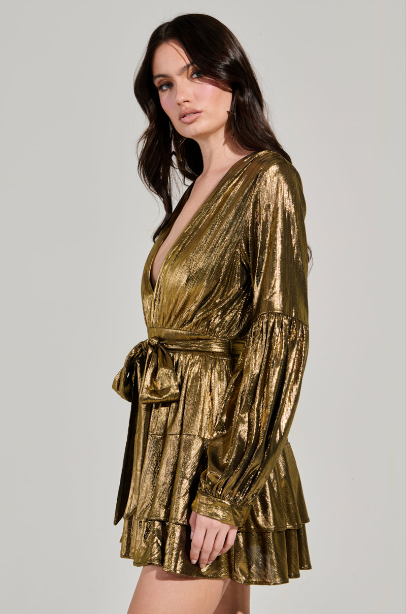 A woman with long, dark hair models the GLOW BABY METALLIC MINI DRESS, showcasing its plunging V-neckline and tiered ruffled skirt. The metallic gold fabric accentuates her style as she poses against a neutral backdrop, looking slightly to the side.