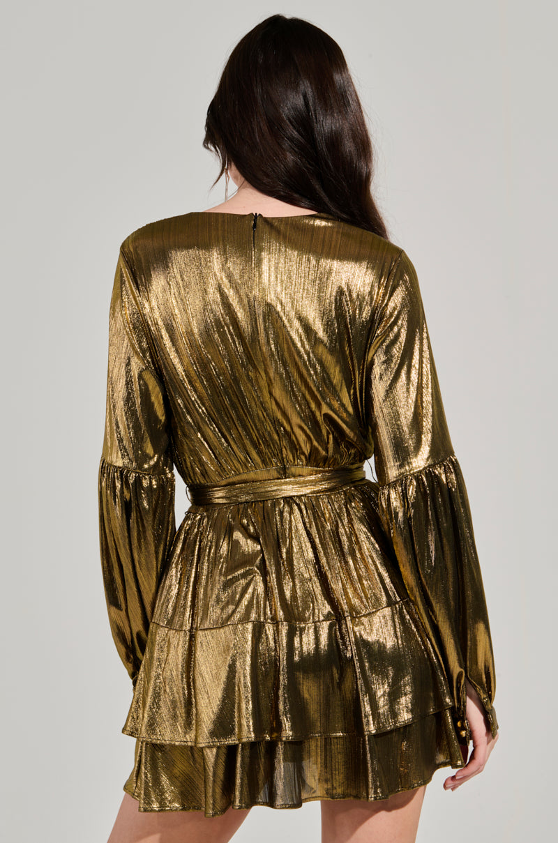 A woman is dressed in the GLOW BABY METALLIC MINI DRESS, showcasing its glistening gold sheen with long sleeves and a tiered ruffled skirt. She poses with her back to the camera, emphasizing the dress's elegant style and shimmering appeal.
