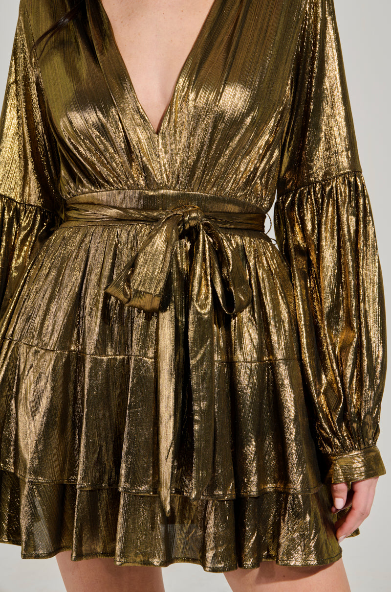 A close-up of a person wearing the GLOW BABY METALLIC MINI DRESS, showcasing its plunging V-neckline and long sleeves. The dress boasts a shimmering gold fabric with a tiered ruffled skirt and a matching belt tied around the waist, highlighting its metallic texture.