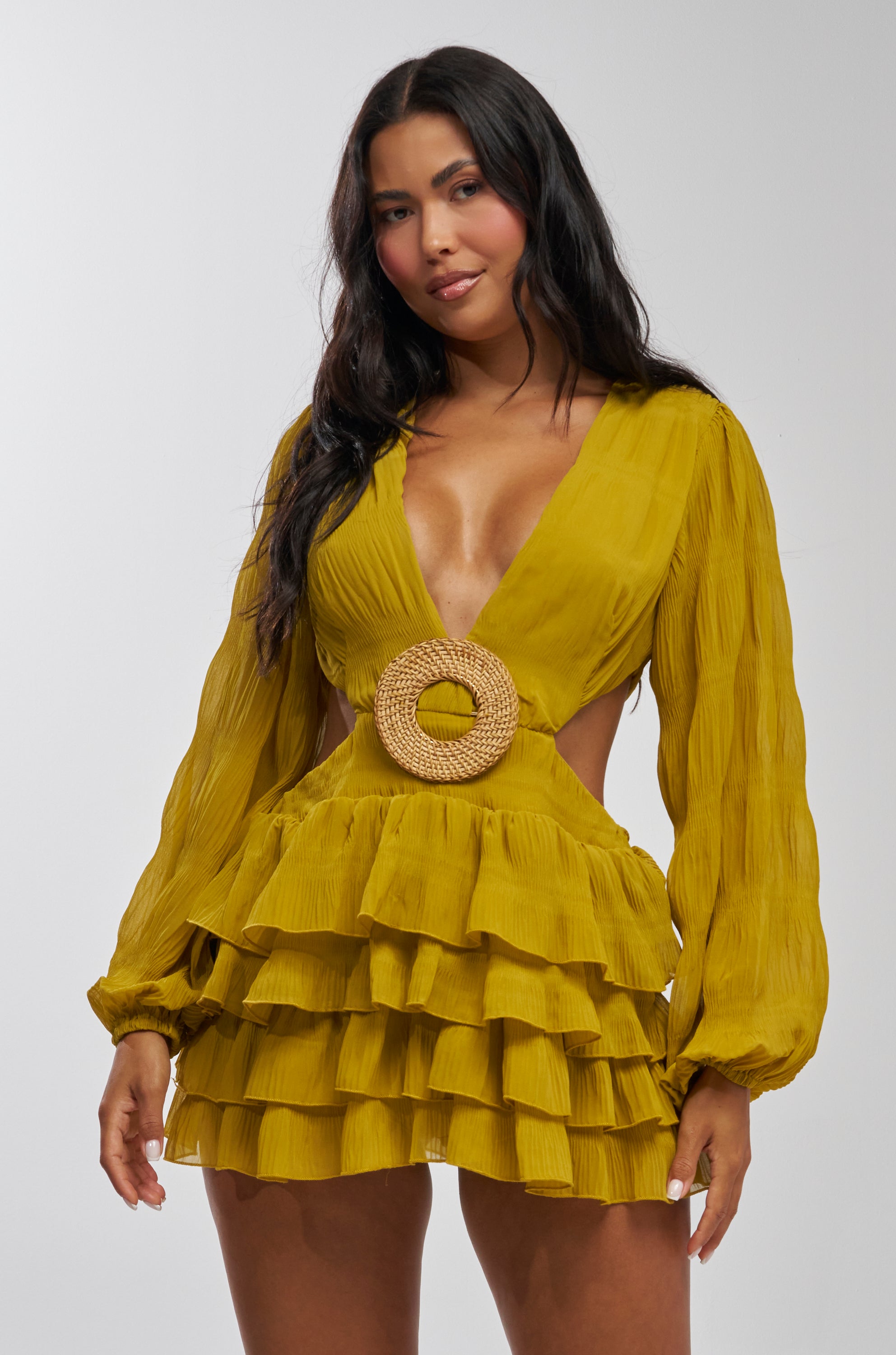A woman with long dark hair models the ON ISLAND TIME RUFFLED MINI DRESS IN CHARTREUSE, featuring ruffled long sleeves, a deep V-neck, circular belt at the waist, side cutouts, and a layered ruffled skirt. She stands against a light background.
