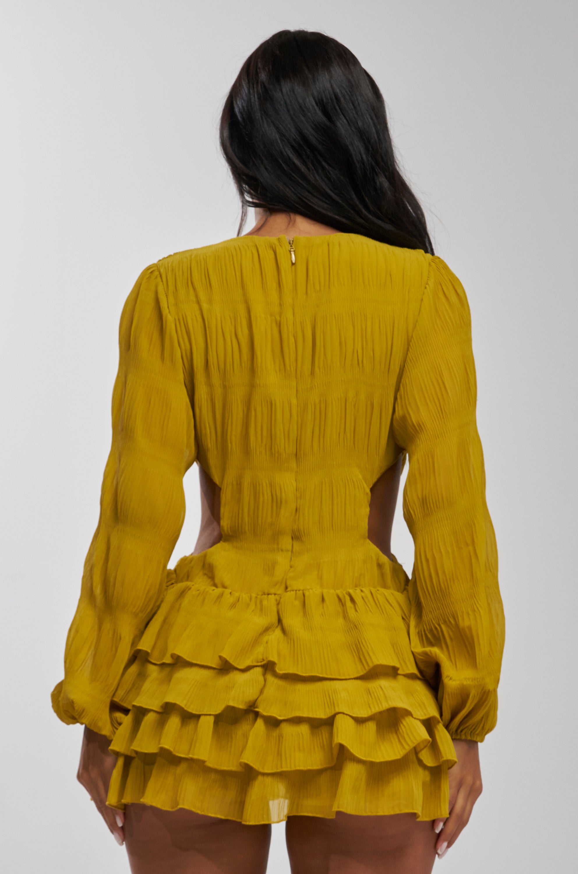A woman with long dark hair is seen from the back, wearing the ON ISLAND TIME RUFFLED MINI DRESS IN CHARTREUSE, which features textured fabric, long sleeves, ruffles, and a layered skirt. The background is plain and light-colored.