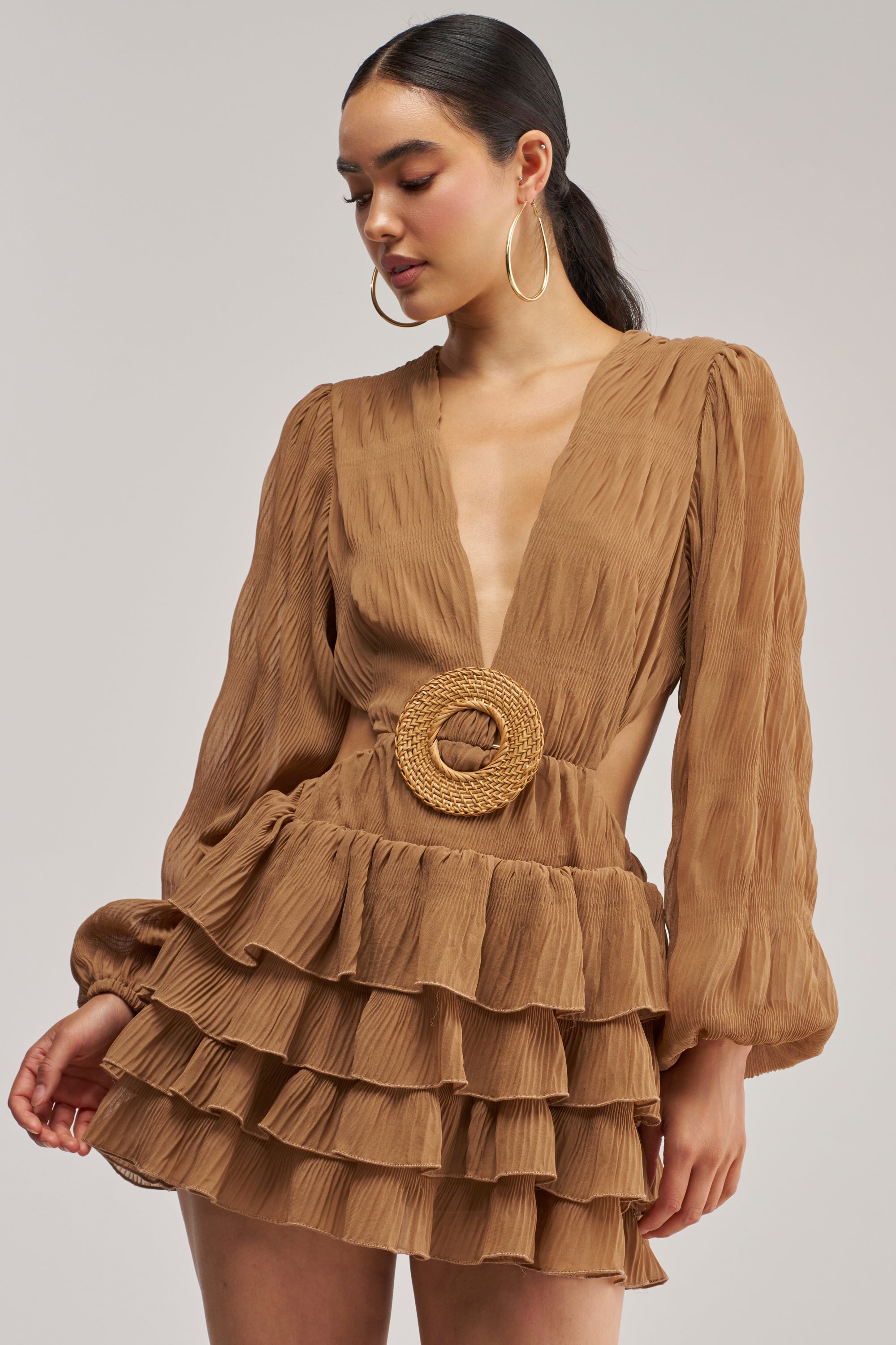 Model wearing the ON ISLAND TIME RUFFLED MINI DRESS IN TAUPE, featuring a deep V-neckline, long sleeves, ruffled tiers, and a circular woven belt at the waist. She completes the look with her hair tied back and large hoop earrings—making it an ideal choice for a summer date night.