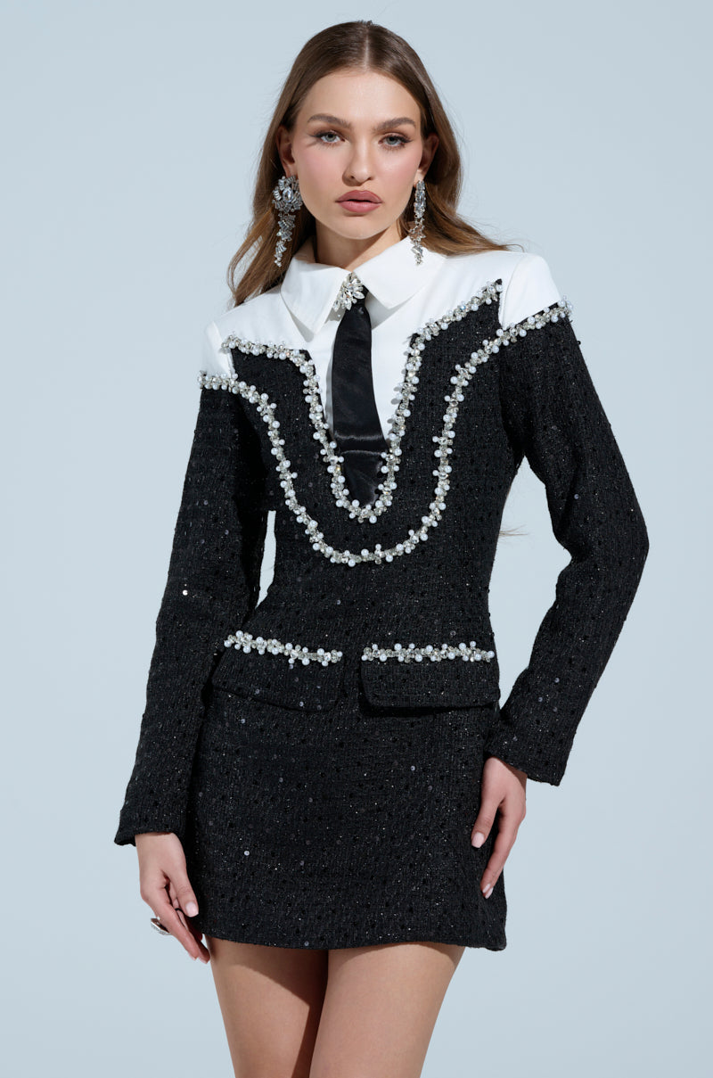 A woman models the High Class Mini Dress, styled with a rhinestone and sequin-embellished jacket over a white collared shirt, black tie, and chic tweed overlay. Her hair is long and paired with drop earrings.