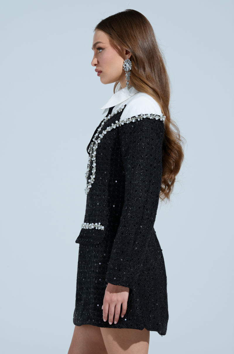 A person with long, wavy brown hair wears a High Class Mini Dress with tweed overlay and rhinestone embellishments. They stand in profile against a plain light blue background, adorned with large dangling earrings complementing their elegant attire.