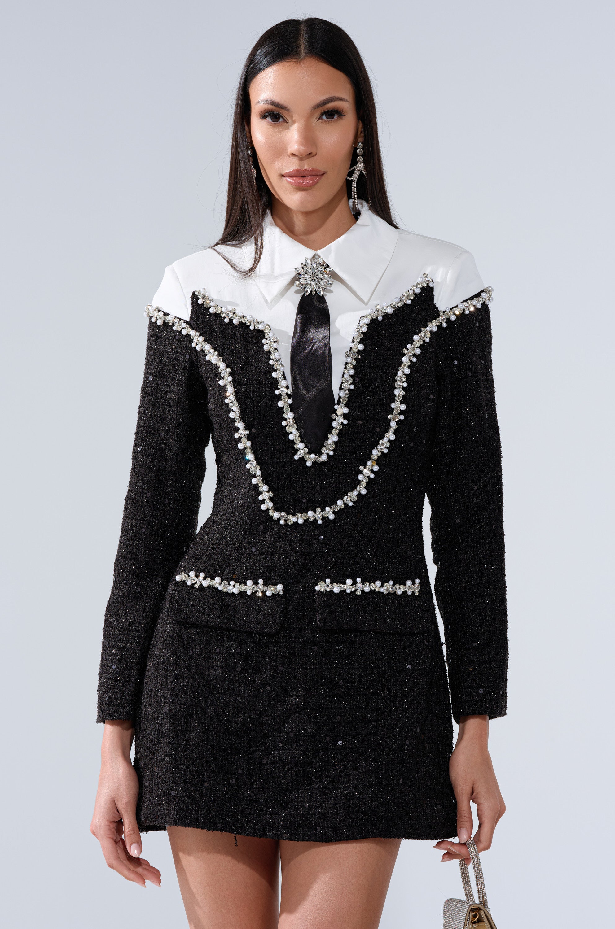 A woman models the HIGH CLASS MINI DRESS WITH A TIE, featuring a black tweed overlay, pearl rhinestone accents, white collared detail, black tie, and statement earrings against a plain light background.