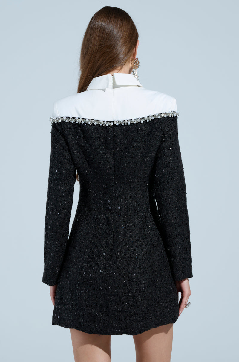 A woman with long brown hair wears the "High Class Mini Dress with a Tie," showcasing its black textured design, jeweled white collar, and rhinestone accents. She faces away against a light blue background, highlighting the outfit's detailed back.