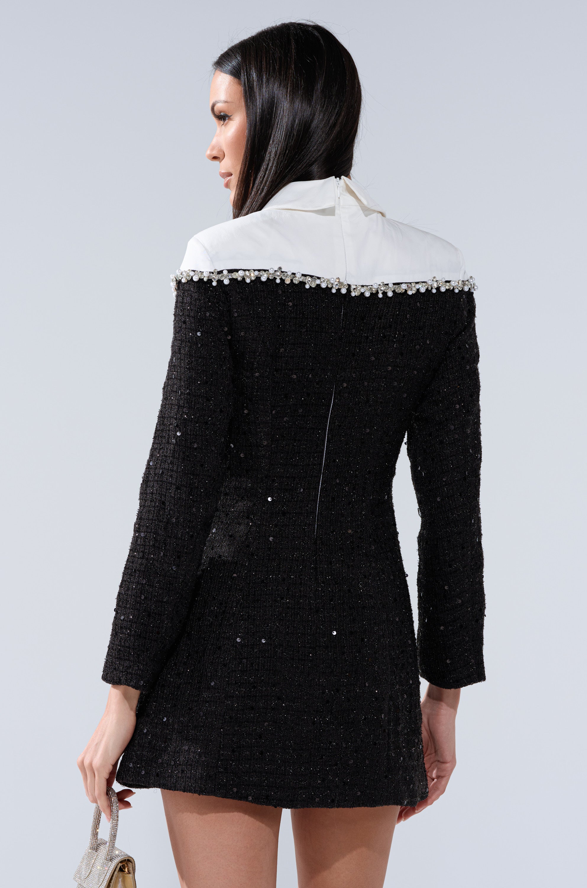 A woman with long dark hair, seen from the back, wears the HIGH CLASS MINI DRESS WITH A TIE featuring black fabric, a white collar, and pearl-trimmed shoulders. She holds a small handbag in her left hand.