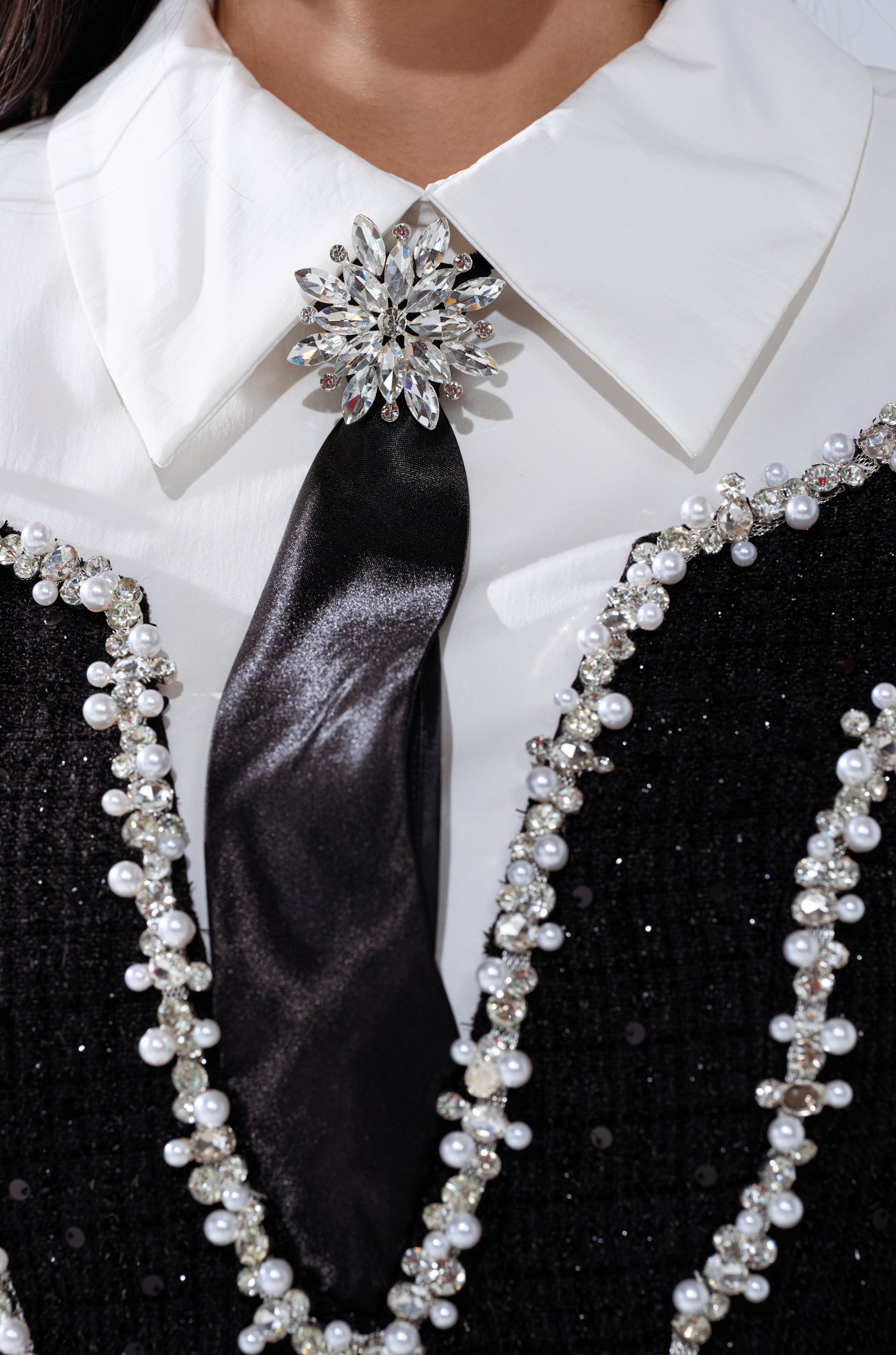A close-up of a person wearing the HIGH CLASS MINI DRESS WITH A TIE—featuring a white collared shirt, black satin tie, crystal brooch, and an embellished mini dress adorned with pearl and bead strands.