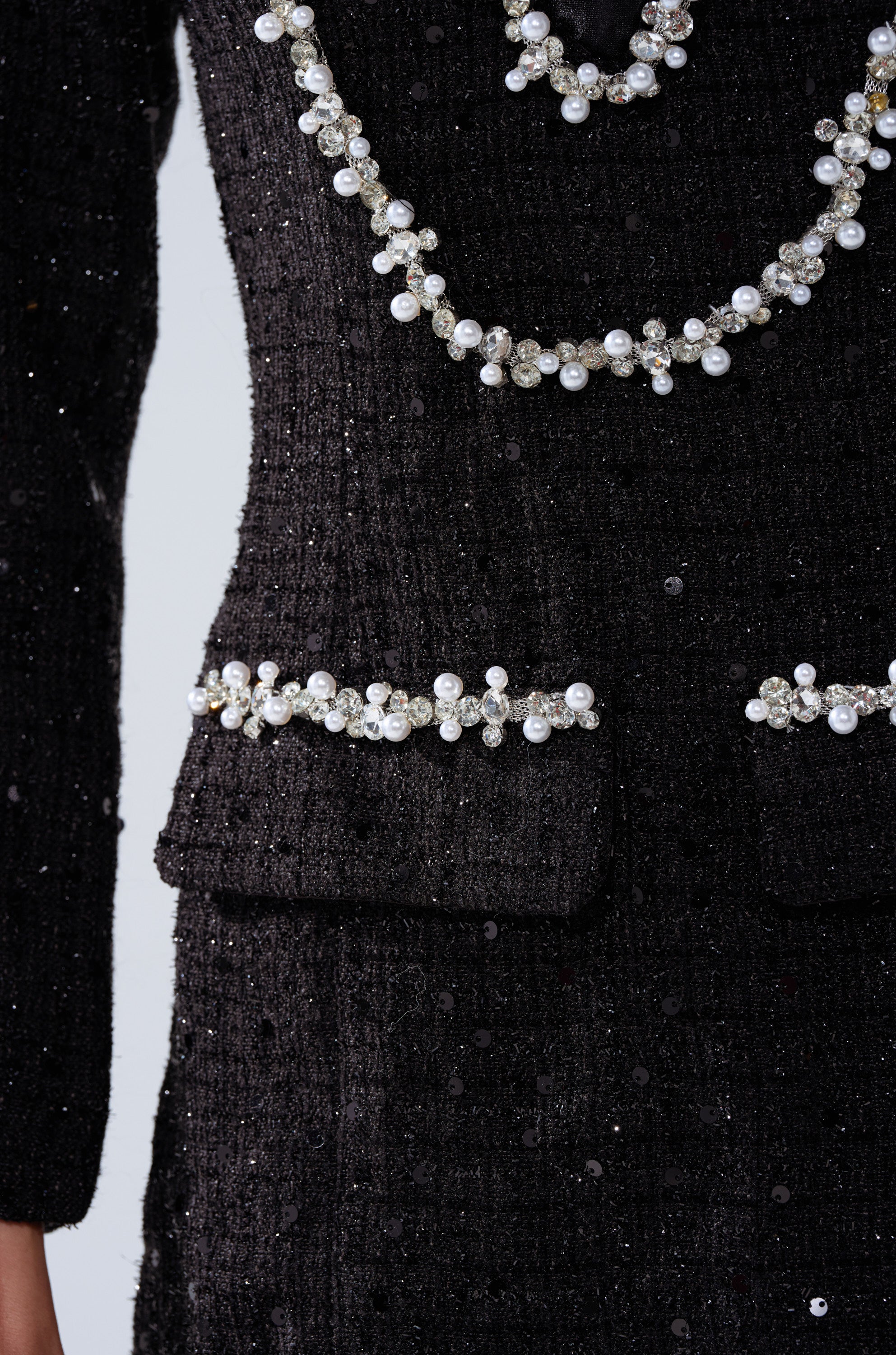 A close-up of the HIGH CLASS MINI DRESS WITH A TIE shows its black tweed, sparkling sequins, and pearl and crystal embellishments on the pockets and necklace. Only the torso and one arm are visible.