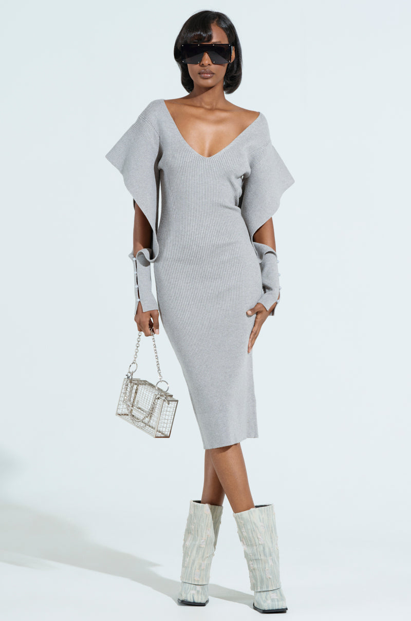 Wearing the "Off to Brunch Knit Midi Dress in Heather Grey," a person stands confidently in a curve-hugging silhouette featuring flounce sleeves and a plunging V-neck. Accessorized with sunglasses, a geometric handbag, and textured high boots, they stand against a plain light background.