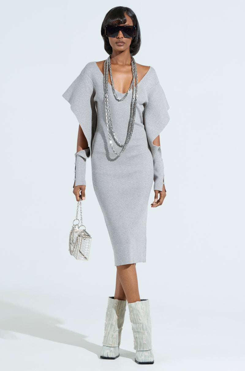 A person wears the OFF TO BRUNCH KNIT MIDI DRESS IN HEATHER GREY with open shoulders and arm cutouts. Accessorized with layered necklaces, large sunglasses, a small handbag, and white textured boots. The curve-hugging look shines against a plain, light-colored background.